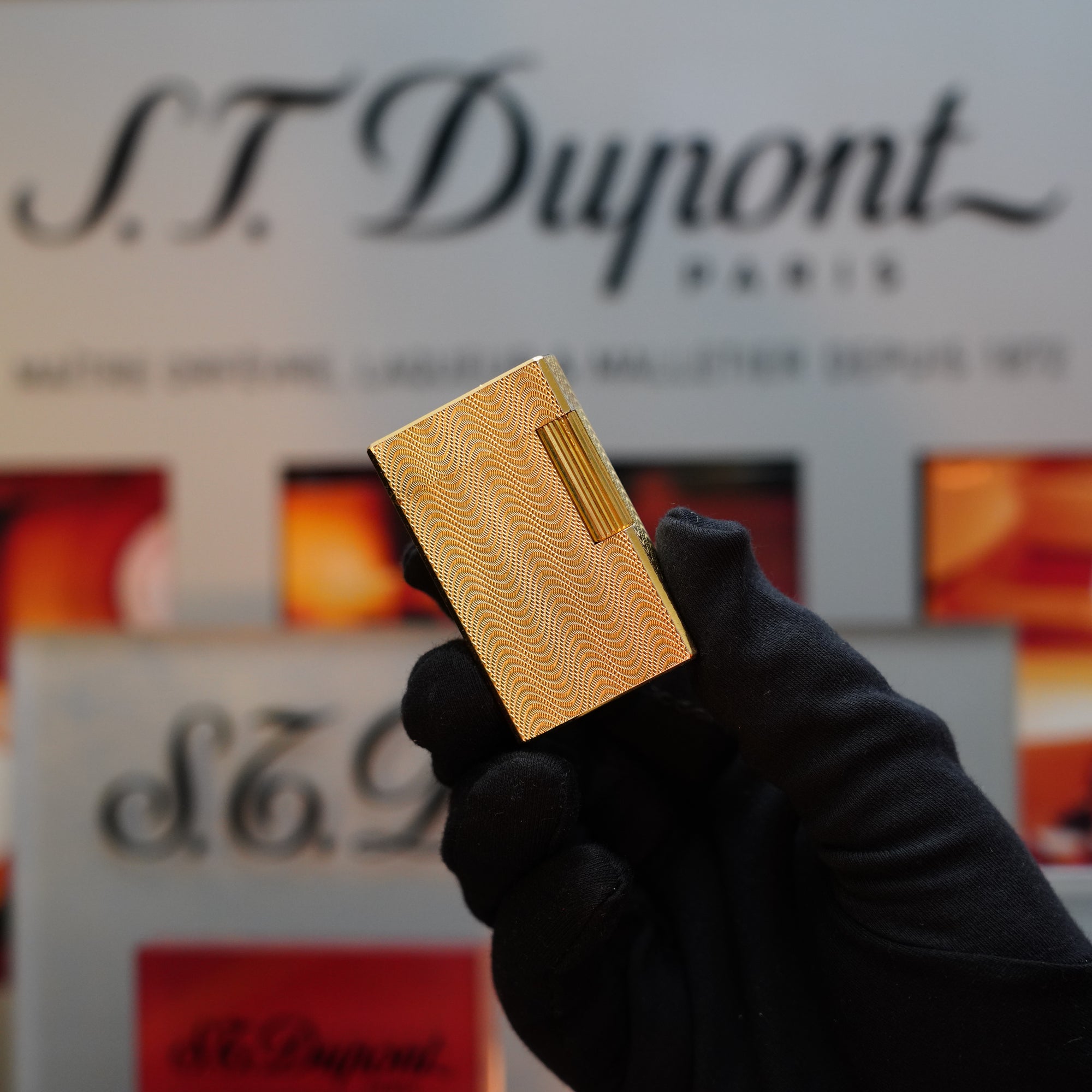 A hand wearing a black glove holds a gold 1960 HERMES Lighter St Dupont, showcasing its exquisite Guilloché pattern reminiscent of the 1960s. The S.T. Dupont signage is visible in the background.