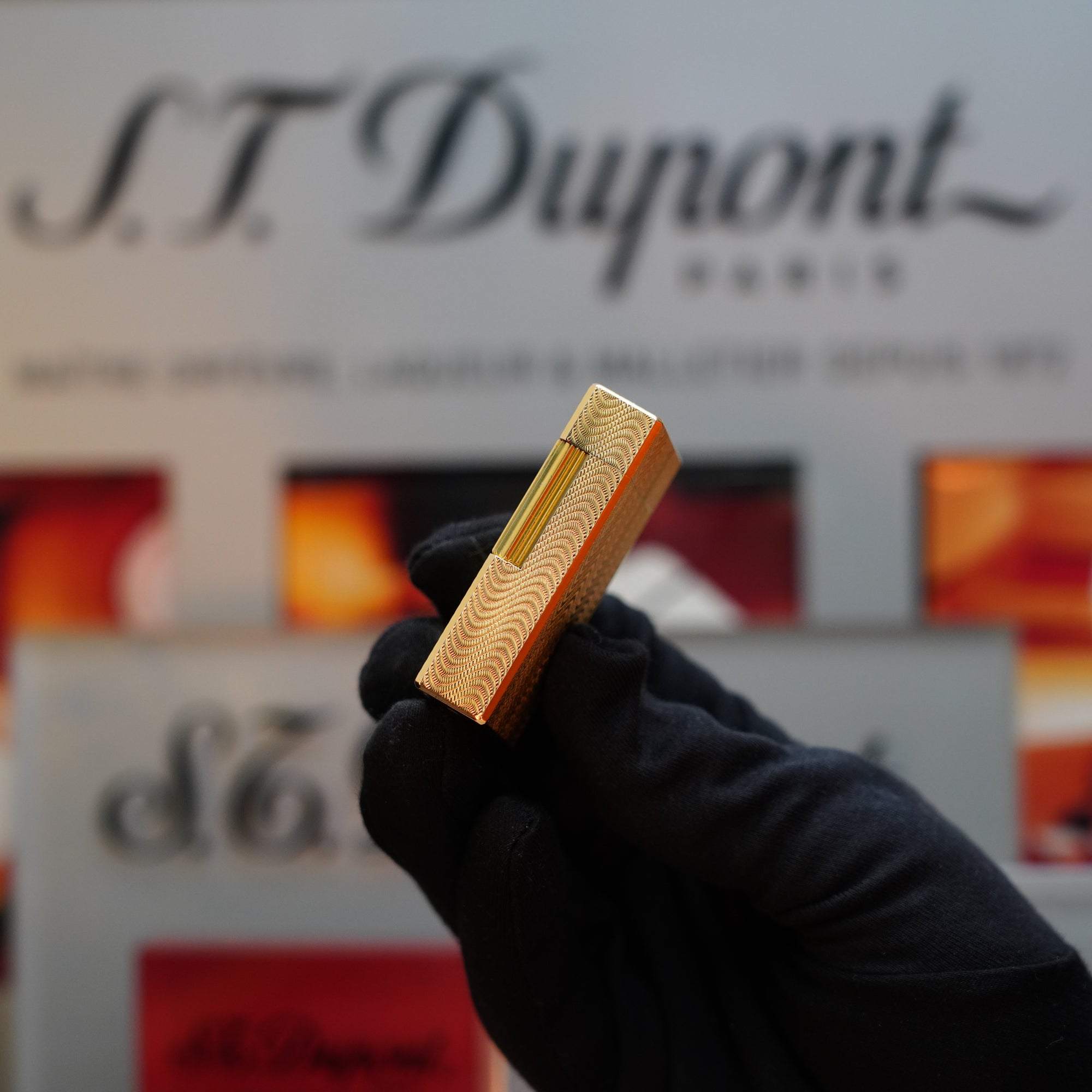 A gloved hand holds a 1960 HERMES Lighter by S.T. Dupont, adorned with a gold finish and intricate Guilloché pattern, in front of a blurred background showcasing S.T. Dupont branding.