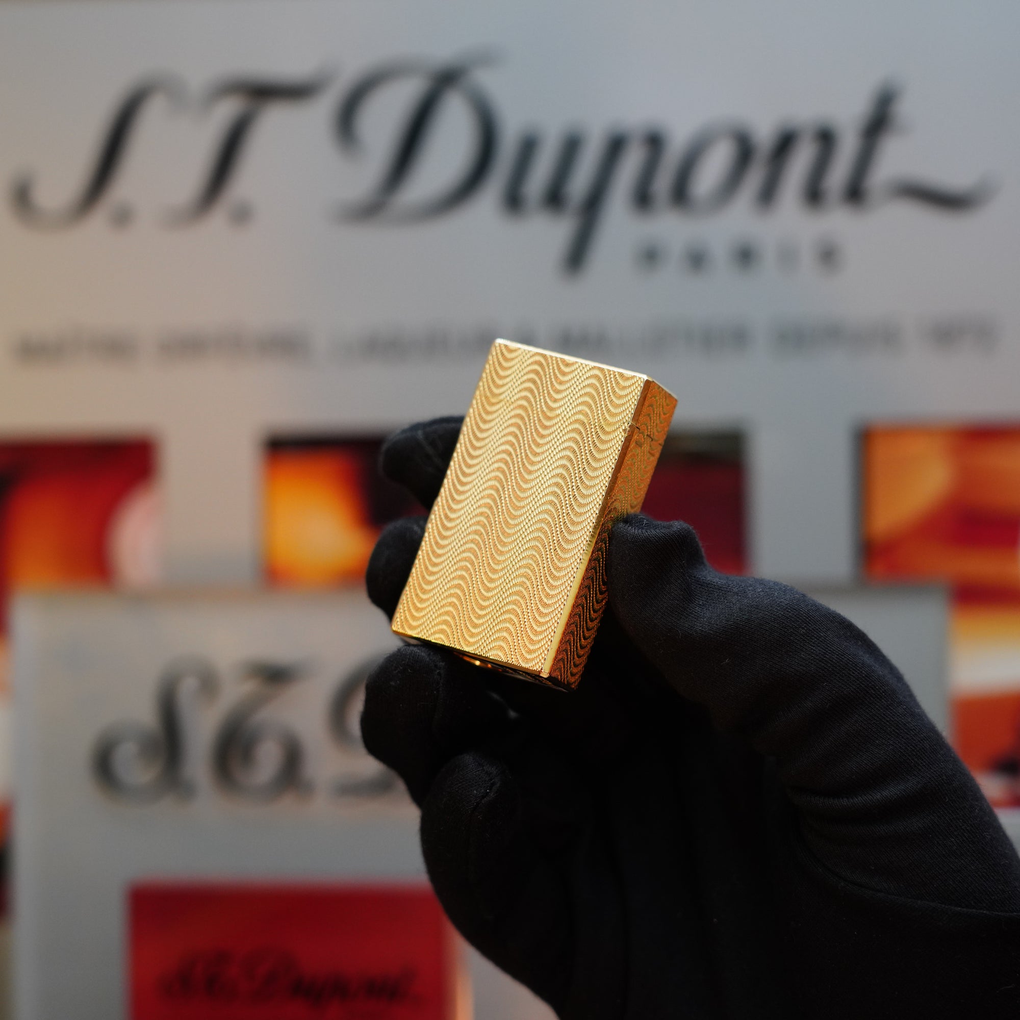 A gloved hand holds the 1960 HERMES Lighter St Dupont Gold Finish Guilloché pattern in front of a blurred S.T. Dupont Paris sign, evoking the elegance of vintage lighters from that era.