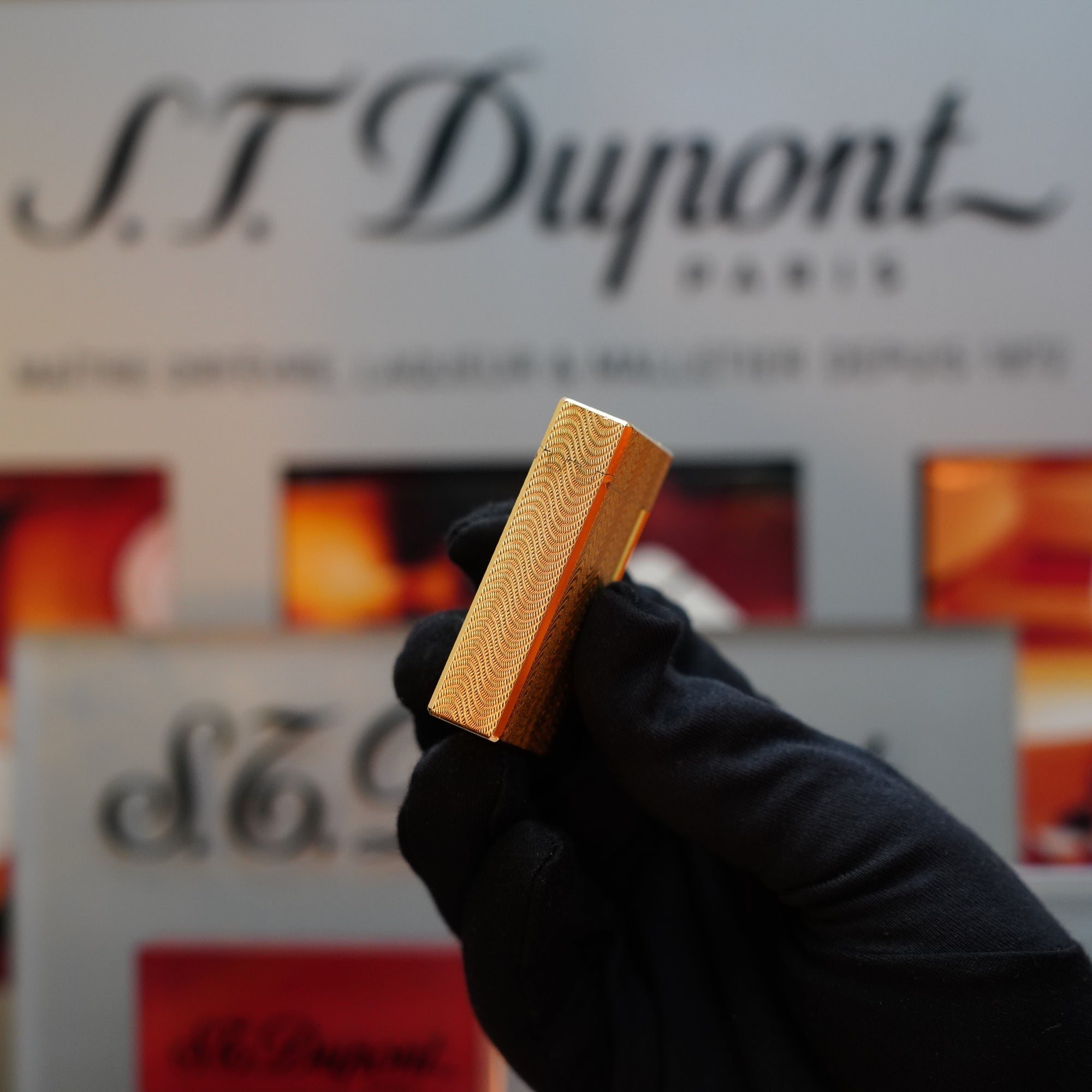 A gloved hand holds a 1960 HERMES Lighter St Dupont with a gold finish and intricate Guilloché patterns against a blurred background featuring the S.T. Dupont Paris logo, reminiscent of vintage elegance.