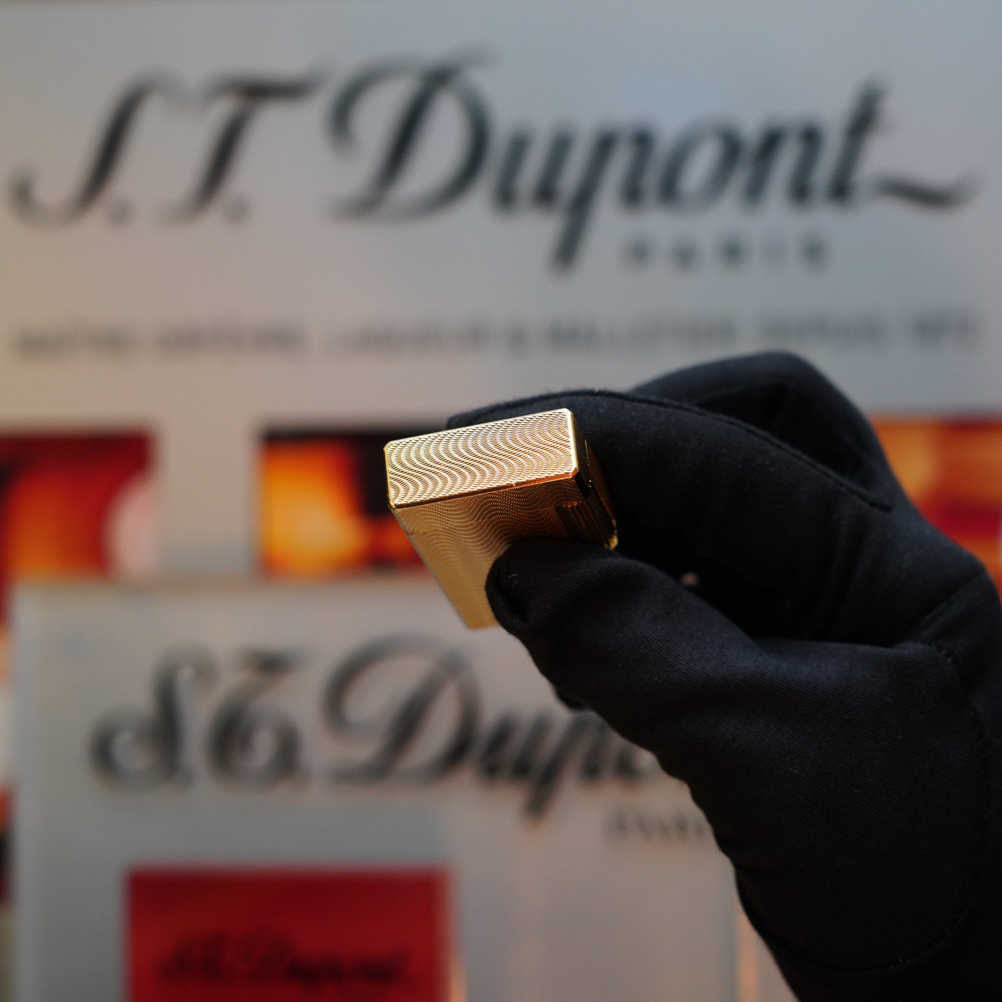 A gloved hand holds a 1960 HERMES St Dupont lighter vertically, its gold finish surface showcasing an intricate Guilloché pattern. The backdrop features a blurred S.T. Dupont sign with red and orange accents, evoking a touch of 1960s elegance.