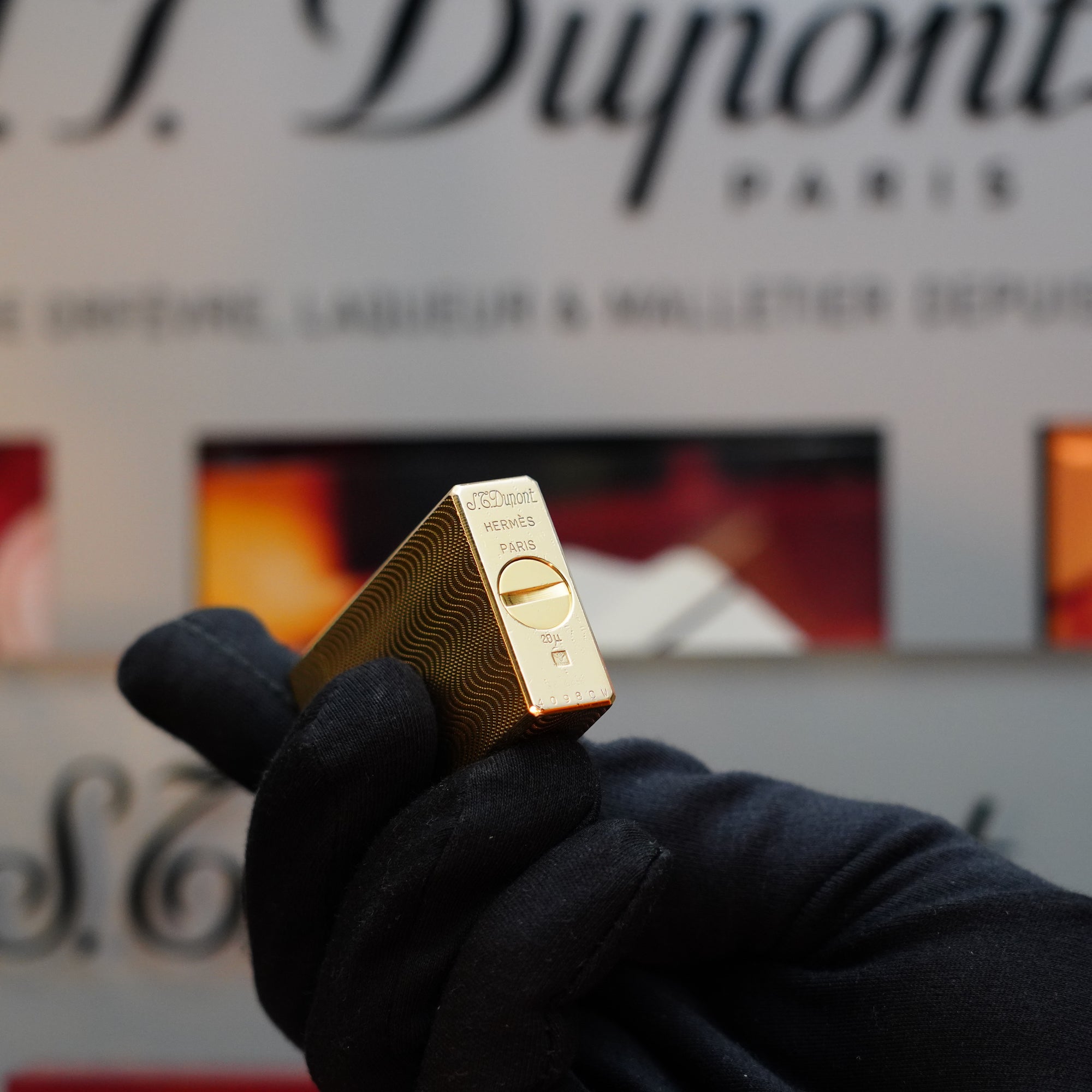 A black-gloved hand holds a 1960 HERMES lighter by S.T. Dupont, angled to showcase its gold finish and intricate guilloché pattern, with the brand name and Paris clearly engraved on it. The background is blurred, leaving "S.T. Dupont" partially visible and evoking the refined elegance of a vintage piece.