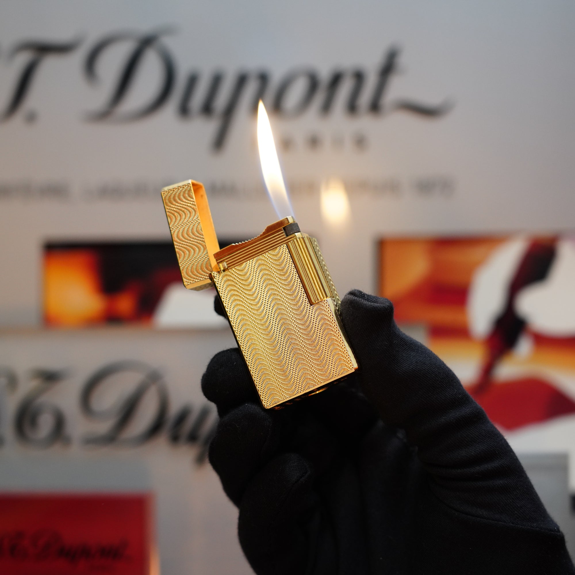 A gloved hand holds a lit 1960 HERMES Lighter St Dupont Gold Finish with a Guilloché pattern against a blurred background, adorned with the "S.T. Dupont" logo.