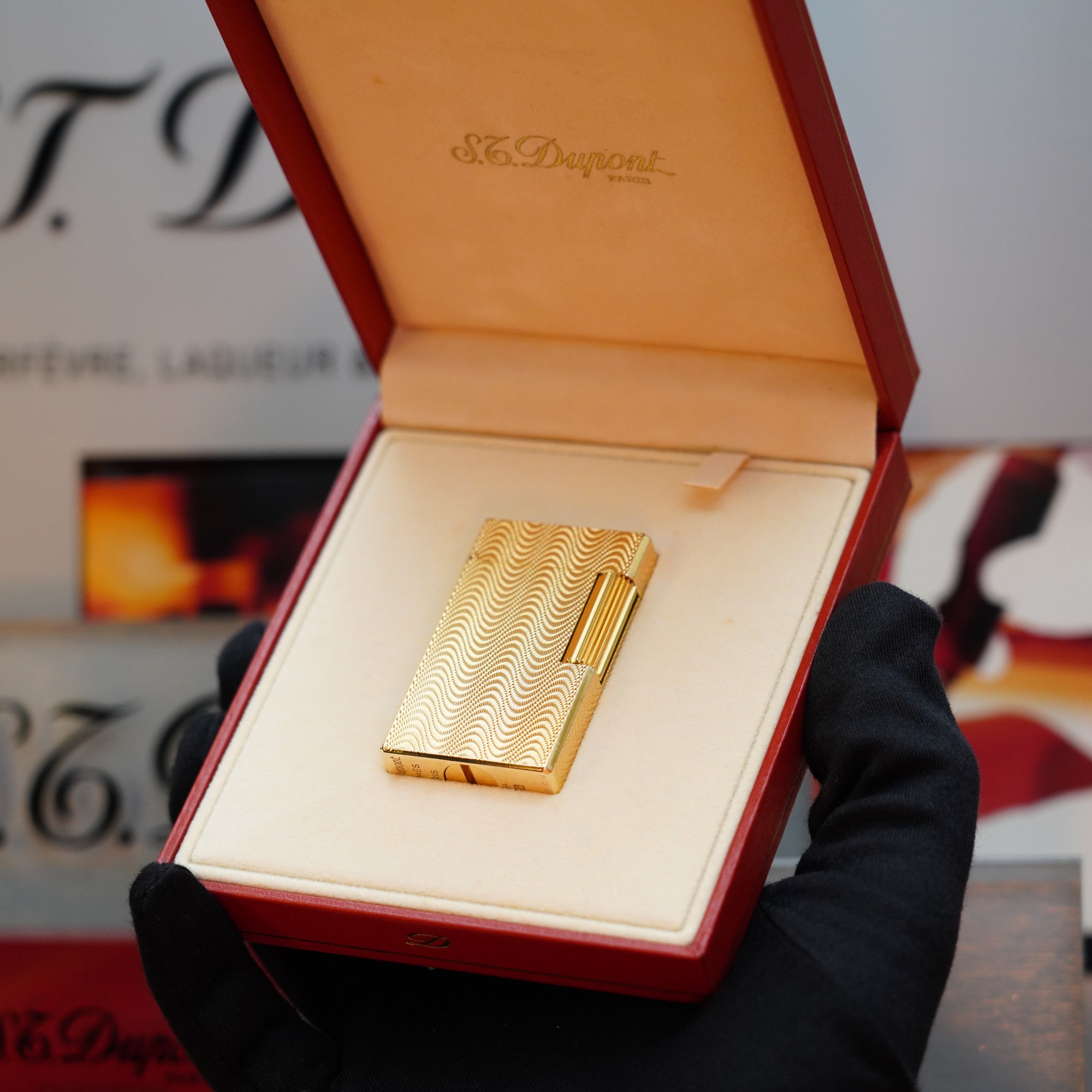 A person wearing black gloves holds an open red box displaying a gold-finished, 1960 HERMES S.T. Dupont lighter with a Guilloché pattern inside.