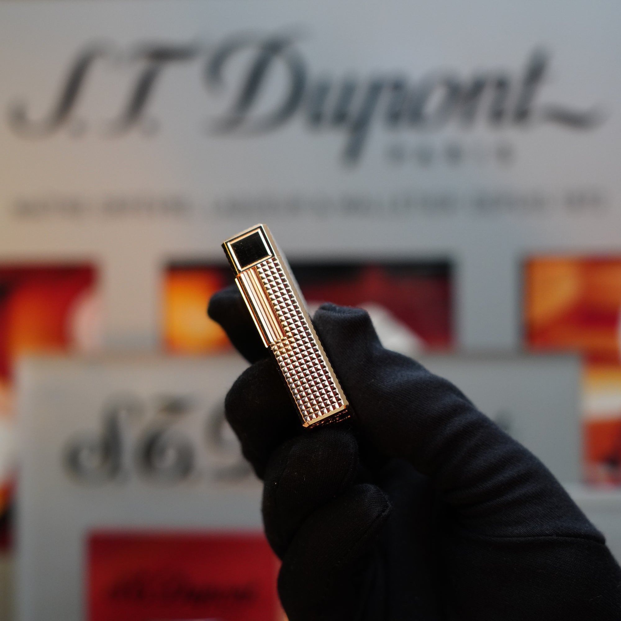 A gloved hand holds a vintage 1960 St Dupont BS lighter from S.T. Dupont, showcasing a luxurious 24k gold finish with a diamond head pattern. The background features blurred S.T. Dupont branding.