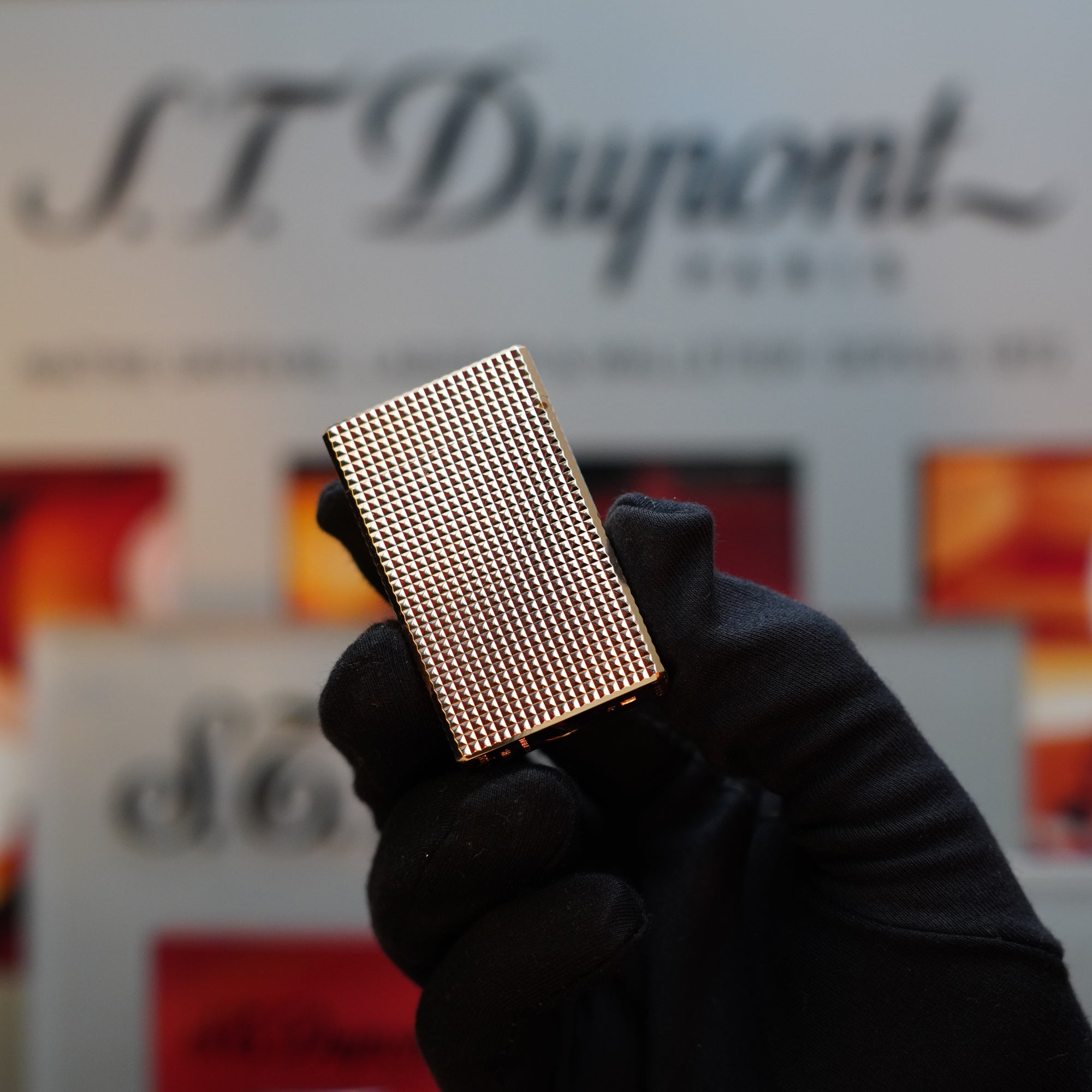 A gloved hand holding a gold-textured rectangular object with an "S.T. Dupont" sign in the blurred background, reminiscent of a vintage 1960 St Dupont BS 24k Diamond Head Gold Finish Rare Set from S.T. Dupont.