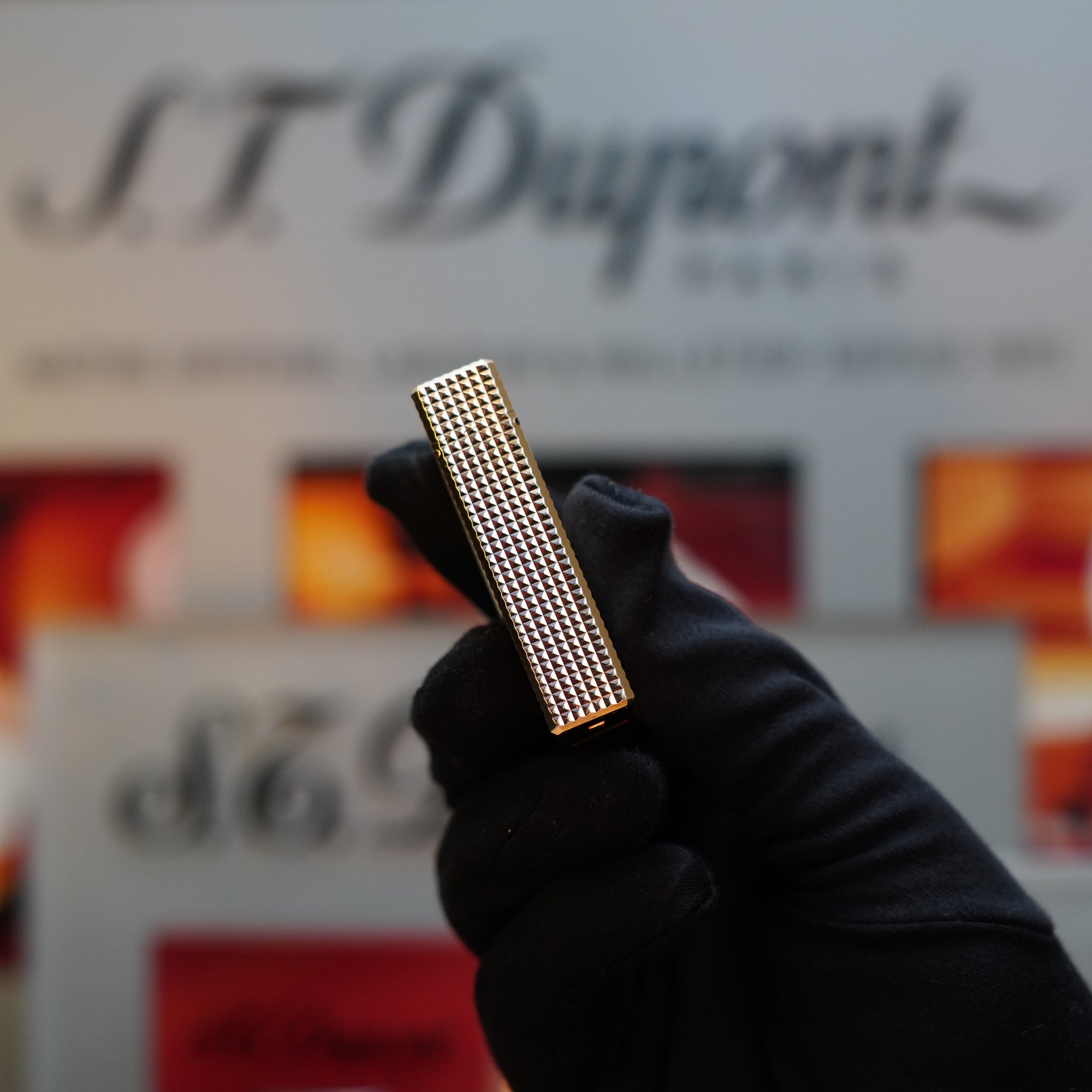 A gloved hand holds a silver-textured 1960 St Dupont BS 24k Diamond Head Gold Finish Rare Set lighter, its vintage charm accentuated by a sleek 24k gold finish, in front of a blurred background featuring the branding and products of S.T. Dupont.