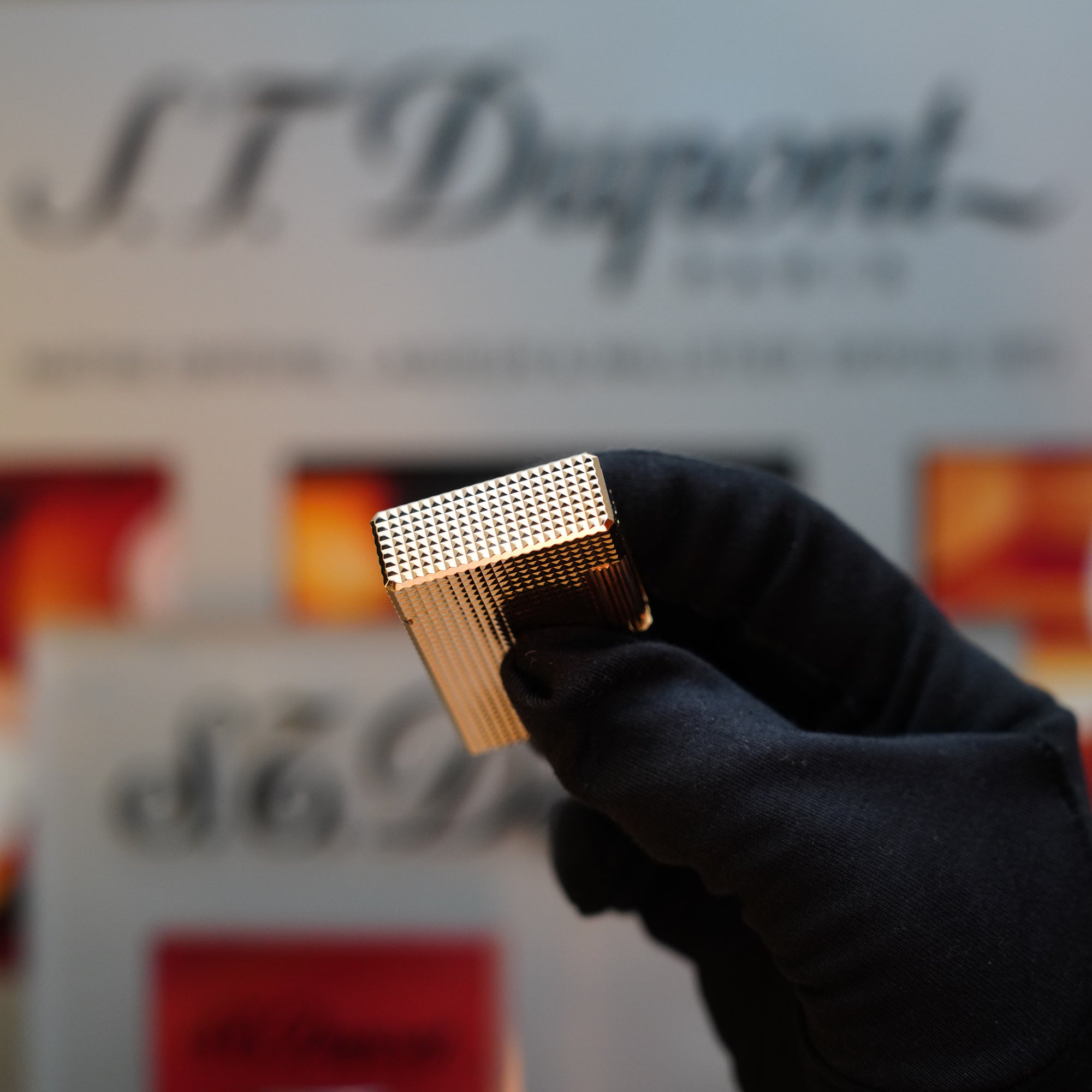 A gloved hand holds a gold-colored, textured rectangular object in front of a blurred background with "S.T. Dupont" branding, reminiscent of the 1960 St Dupont BS 24k Diamond Head Gold Finish Rare Set from S.T. Dupont.