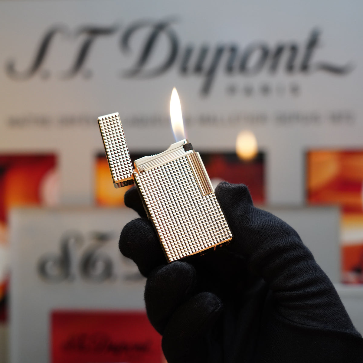 A gloved hand holds a lit 1960 St Dupont BS 24k Diamond Head Gold Finish lighter from the rare set, in front of a blurred S.T. Dupont Paris sign.