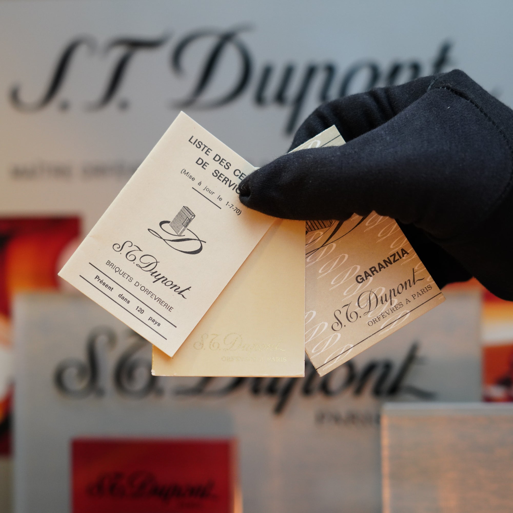 A gloved hand holds two S.T. Dupont guarantee and service list cards against a blurred background featuring the S.T. Dupont brand name, reminiscent of the elegance found in their 1960 St Dupont BS 24k Diamond Head Gold Finish Rare Set.