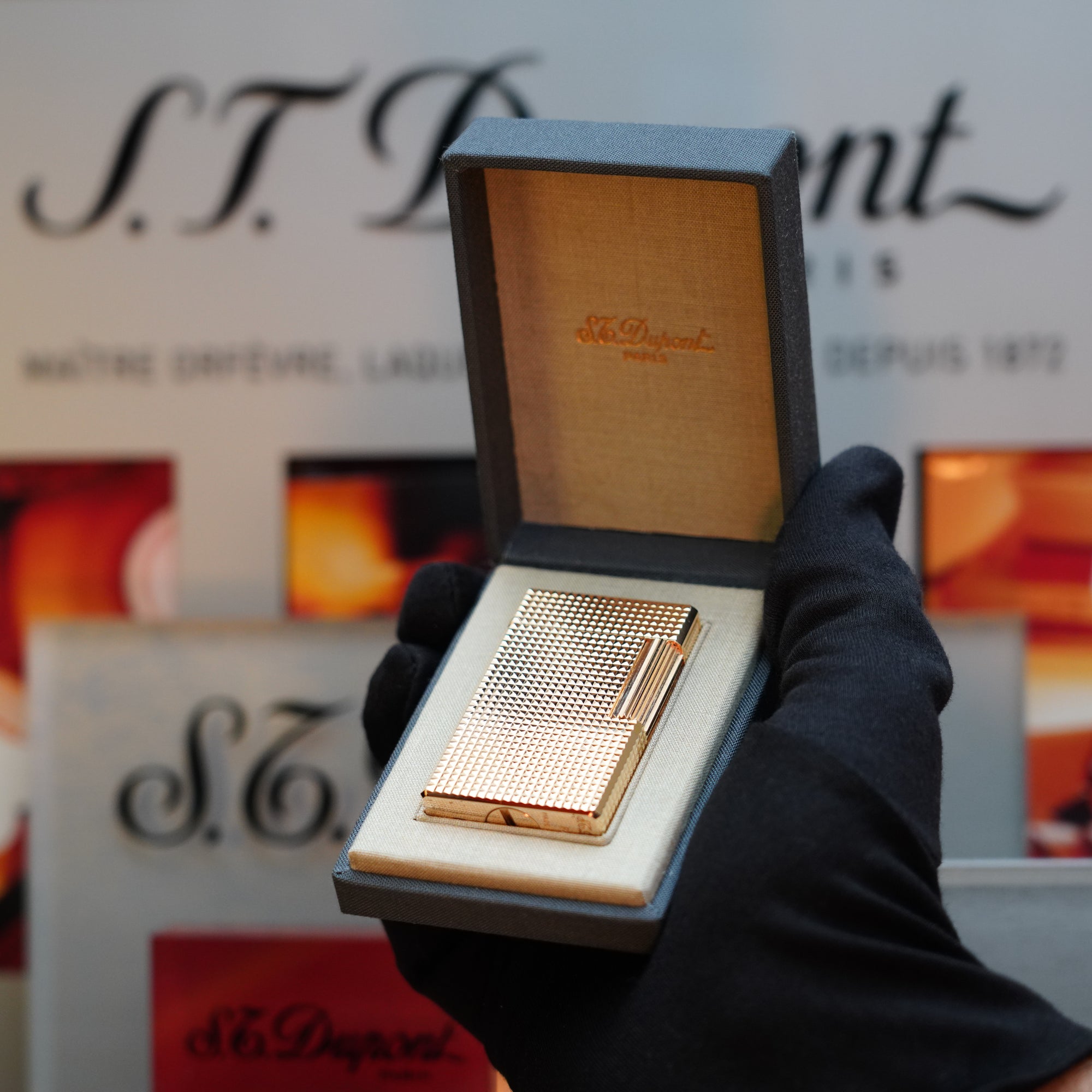 A black-gloved hand holds an open box with a 1960 S.T. Dupont BS 24k Diamond Head Gold Finish lighter inside, glowing with its exquisite gold exterior. In the background is a sign displaying the S.T. Dupont logo and text, evoking the elegance of the vintage Ligne 1 Series collection.