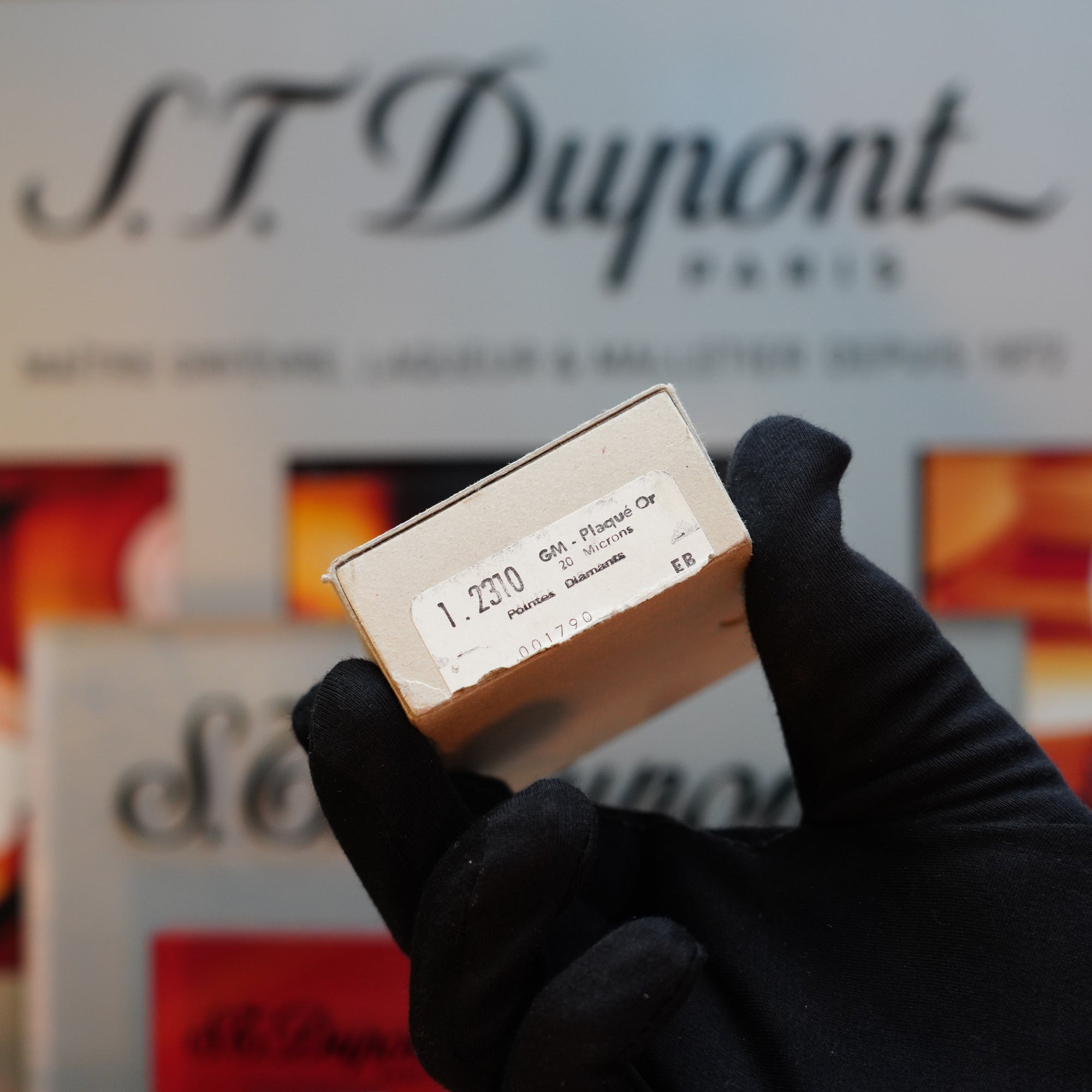 Close-up of a gloved hand holding a small box with a label that reads "G.M. Pionni Or" in front of blurred S.T. Dupont branding, showcasing the elegant 24k gold finish of the 1960 St Dupont BS 24k Diamond Head Gold Finish Rare Set.