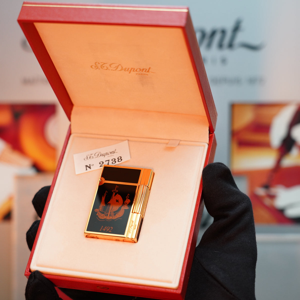 A gloved hand displays the Vintage 1992 S.T. Dupont Important 24k Gold Finish Colombus Gatsby Second Limited Edition Lighter, showcasing its luxurious gold and black finish with a ship design, in an open red velvet box.