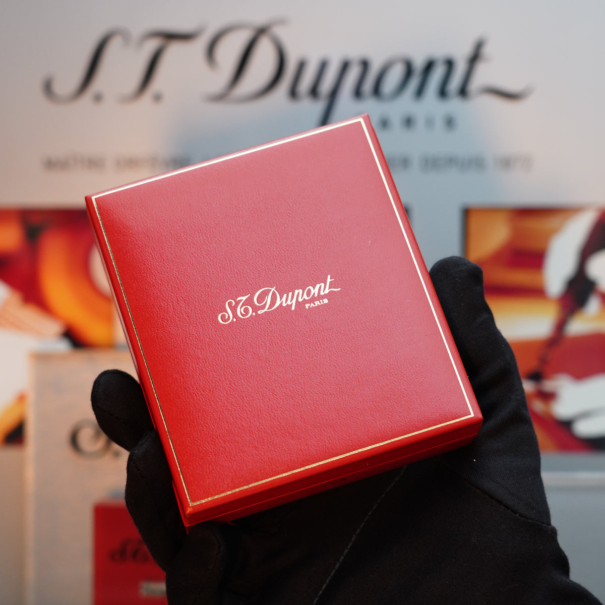 A person wearing a black glove holds a red S.T. Dupont Paris box, hinting at the elegance within—a Vintage 1980 St Dupont Heavy Silver Finish Ligne 1 Fine Hobnail Pattern Grain D`Orge Ecusson Lighter. The brand name stands prominently in the background, reflecting timeless sophistication.
