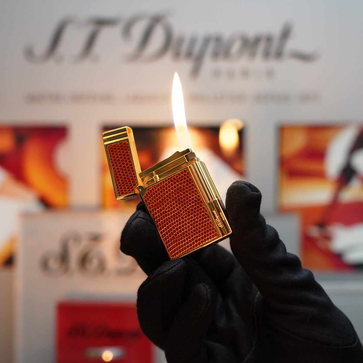 A gloved hand holds a lit Vintage 1980 S.T. Dupont 24k Gold finish Gatsby Gainerie Lezard Brown Authentic leather extremely rare lighter against a blurred background.