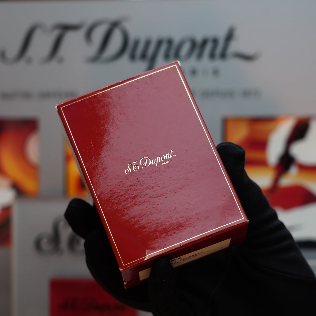 A gloved hand gently holds a red S.T. Dupont Paris box, suggesting the allure of a Vintage 1990 St Dupont Rare Ornee Soubreny 24k Gold Finish Red Passion Lacquer Lighter, as blurred promotional material in the background subtly hints at its mystique.