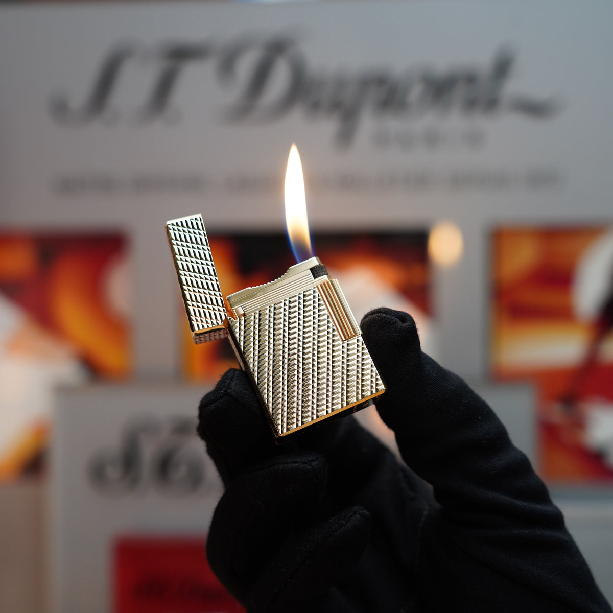 A gloved hand holds an open Vintage 1970 S.T. Dupont Ligne 1 lighter, showcasing its rare visual pattern and heavy 24k Gold Finish with a lit flame, against a blurred background featuring the S.T. Dupont logo.