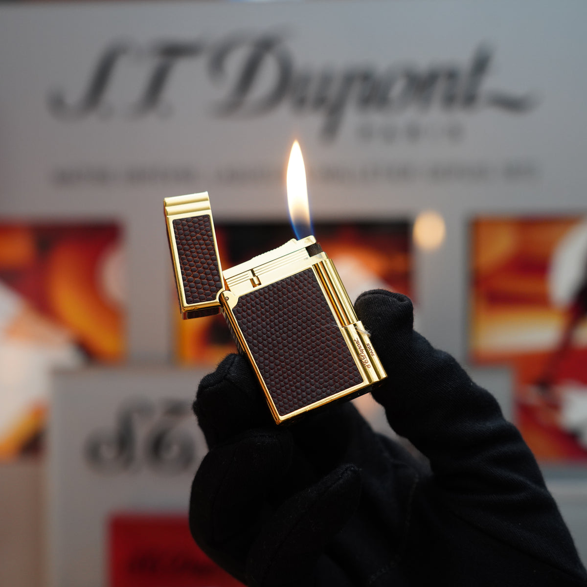 A gloved hand holds an open Vintage 1988 S.T. Dupont lighter showcasing a visible flame, set against a blurred background featuring the brand's logo. The Gatsby Gainerie Lezard Brown Authentic Leather and 24k Gold finish add an exquisite touch to this rare and elegant piece.
