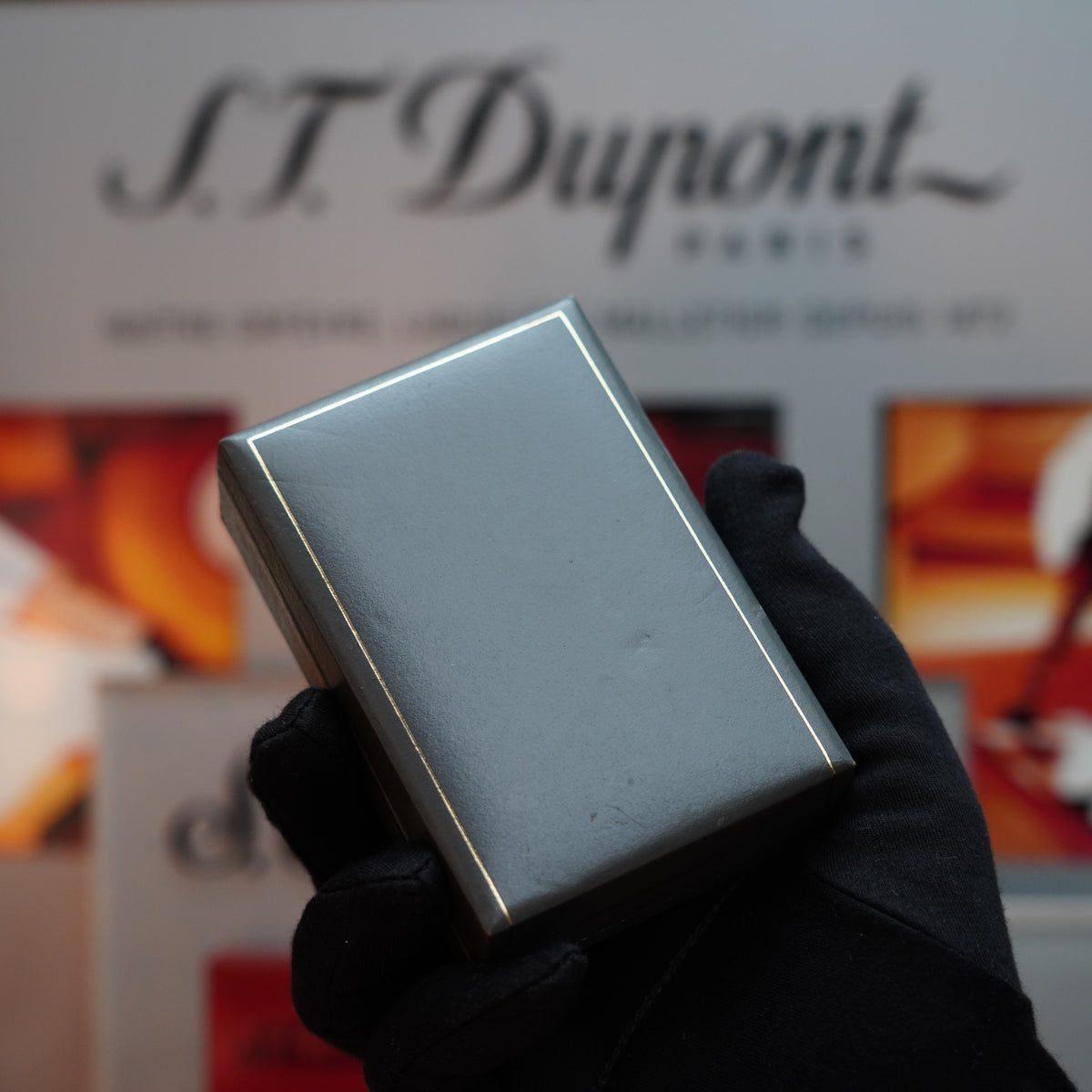 A gloved hand presents a silver rectangular box, potentially housing the rare Vintage 1980 Dunhill D line Rollagas Cross Hatch Heavy Silver Finish lighter, against the backdrop of a blurred S.T. Dupont Paris display, evoking elegance reminiscent of high-end Dunhill craftsmanship.