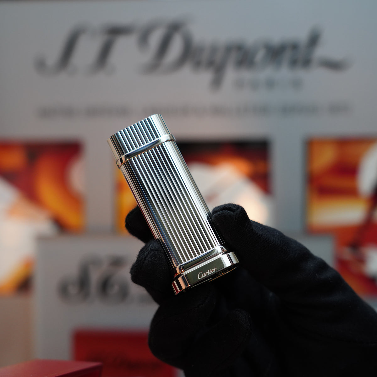 A gloved hand holds a Cartier lighter, the smooth palladium finish of the Vintage 1990 Cartier Pasha Oval Le Must Table Lighter gleaming against the blurred store display background.