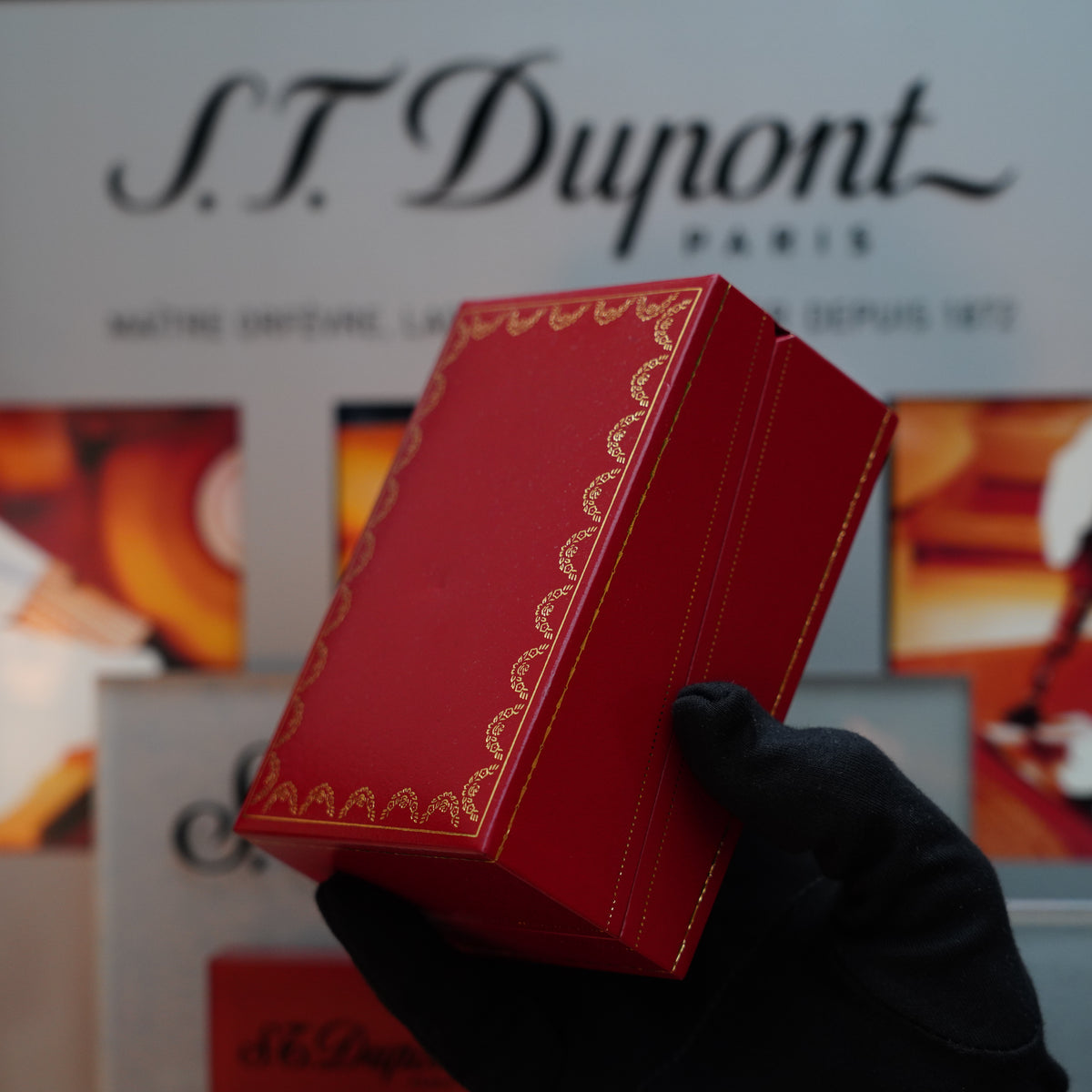 A gloved hand holds a red box with gold detailing, reminiscent of a Vintage 1990 Cartier Pasha Vertical Line Heavy Palladium Finish Oval Le Must Table Lighter, in front of an S.T. Dupont Paris display.