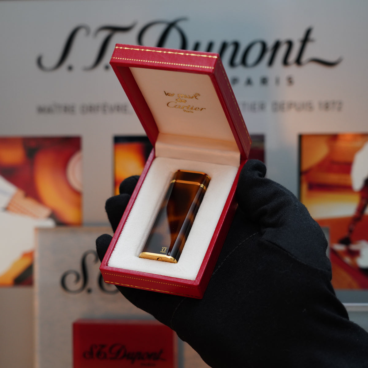 A gloved hand holds a red box containing the Vintage 1980 Cartier Rare Panther 24k Gold Heavy Finish iconic Panther Tortoiseshell Lacquer Oval Le Must Lighter, set against a blurred S.T. Dupont display background.