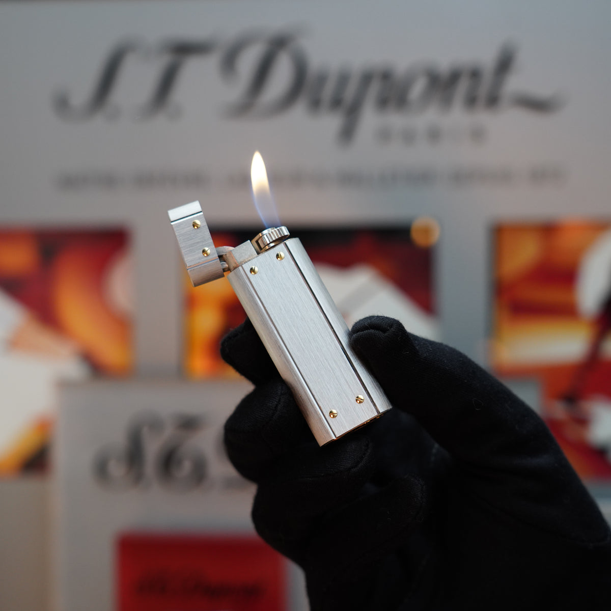 A hand in a black glove elegantly holds a lit Vintage 1980 Cartier Santos lighter, featuring a palladium finish with 24k gold screws, against a blurred background, echoing the ambiance of timeless sophistication and Cartier branding.