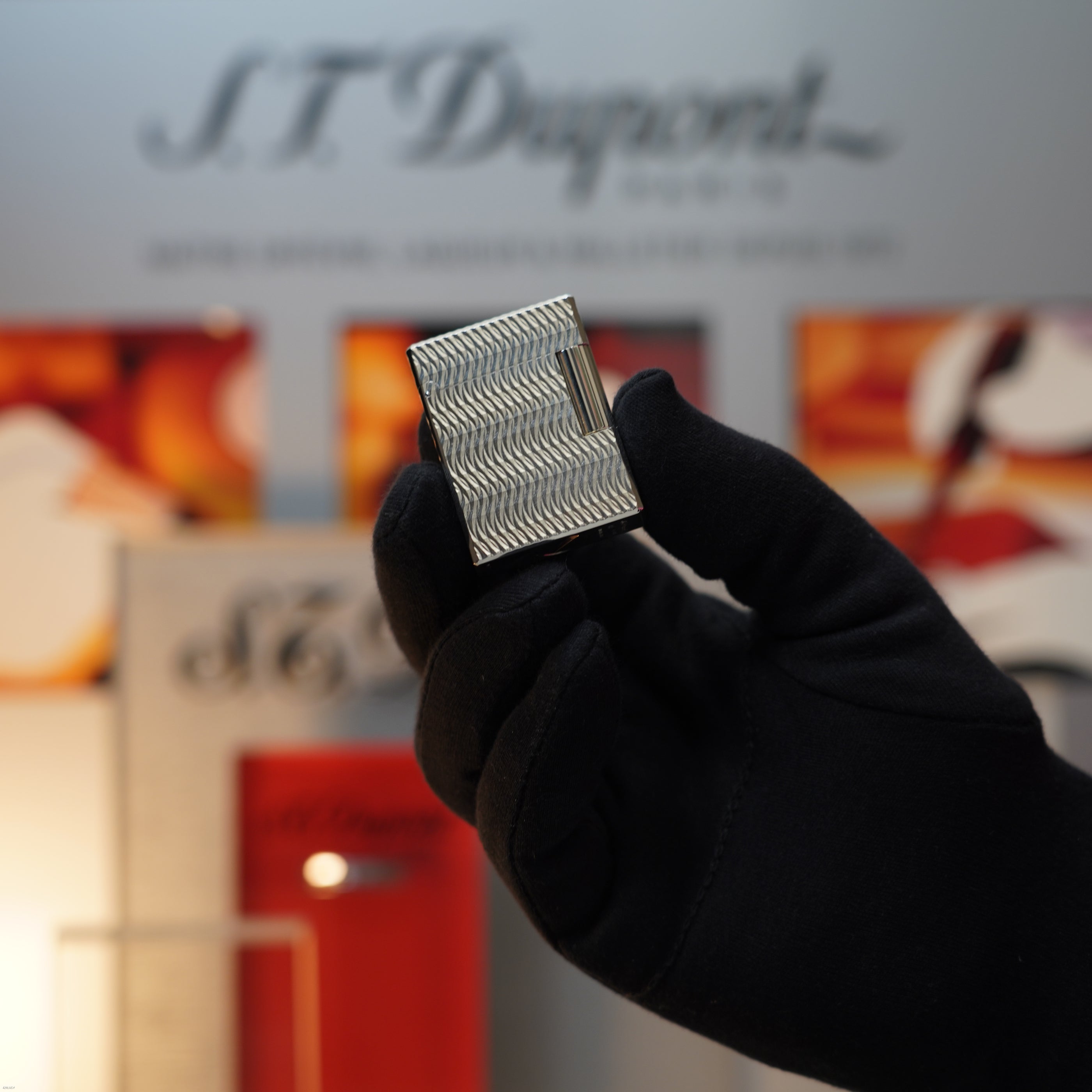 A gloved hand holds a Lucy Lighter Vintage 1990 S.T. Dupont Ligne 1 Small Size Rare 3D Wave Pattern Heavy Silver Finish Lighter, with a blurred S.T. Dupont display in the background.