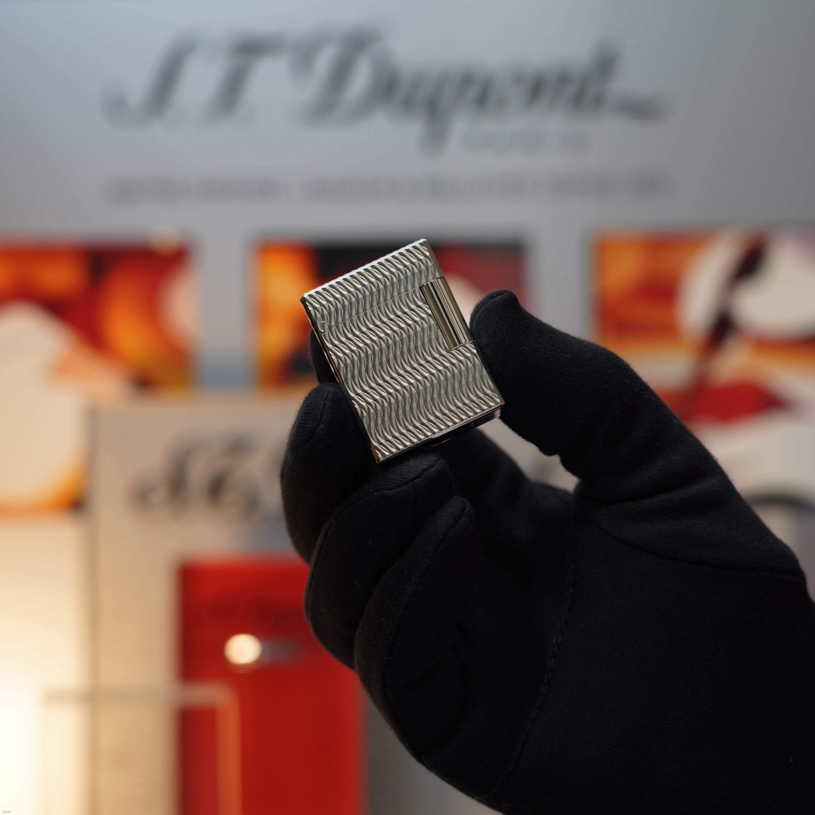 A gloved hand holds a Lucy Lighter Vintage 1990 S.T. Dupont Ligne 1 small-size rare 3D wave pattern heavy silver finish lighter, with an S.T. Dupont display blurred in the background.