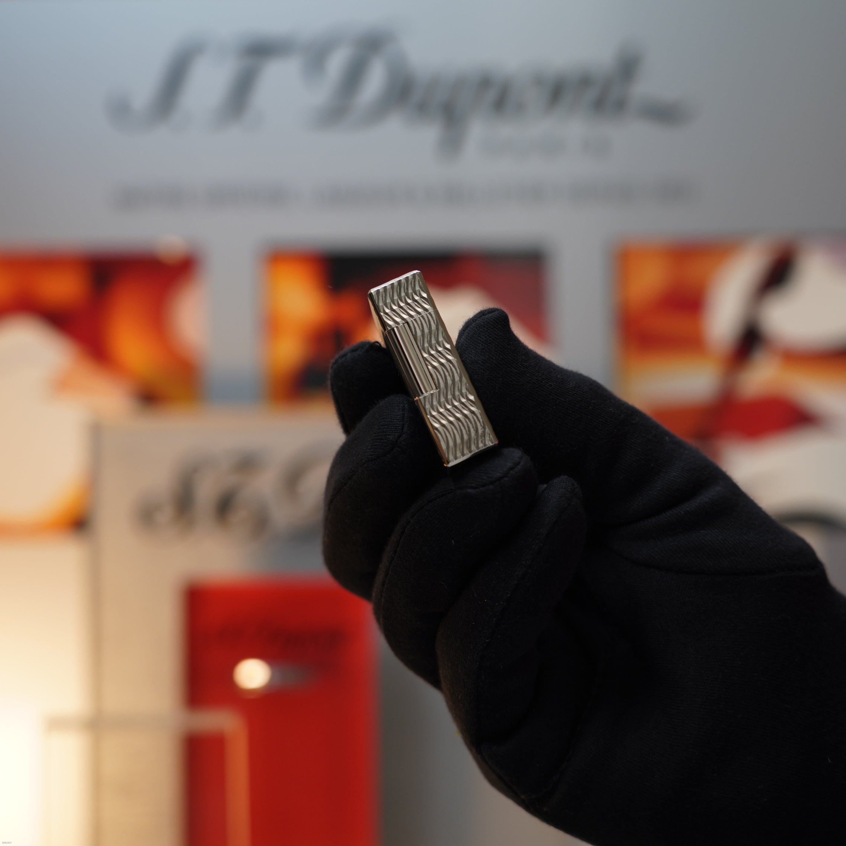 A gloved hand holds a Lucy Lighter Vintage 1990 S.T. Dupont Ligne 1 Small Size Rare 3D Wave Pattern Heavy Silver Finish Lighter in front of a blurred display.