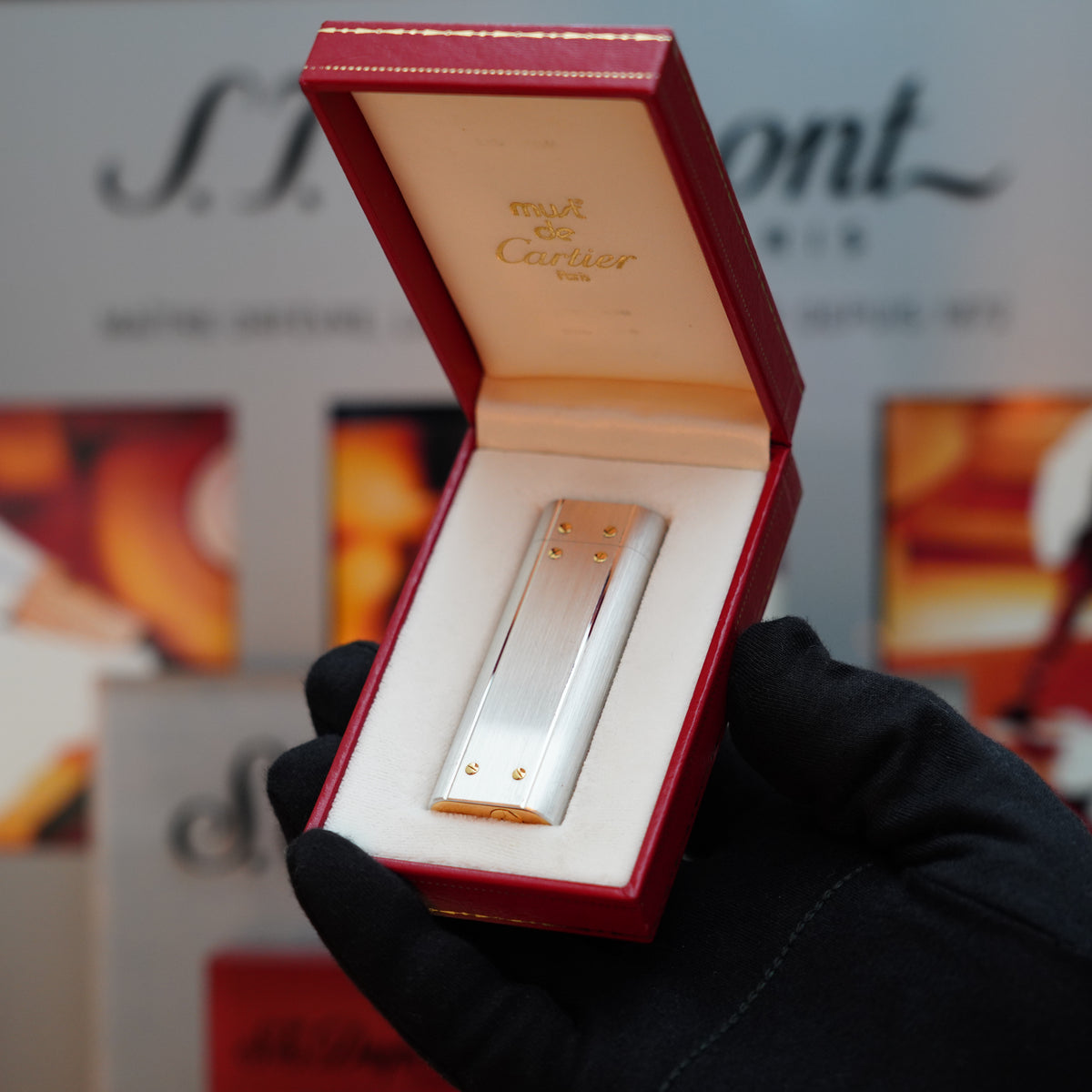 A person wearing a black glove holds an open red Cartier box, unveiling a Vintage 1980 Cartier Santos Le Must Lighter inside—a stunning fusion of palladium and 24k gold finishes that captures the elegance of a classic Cartier lighter.