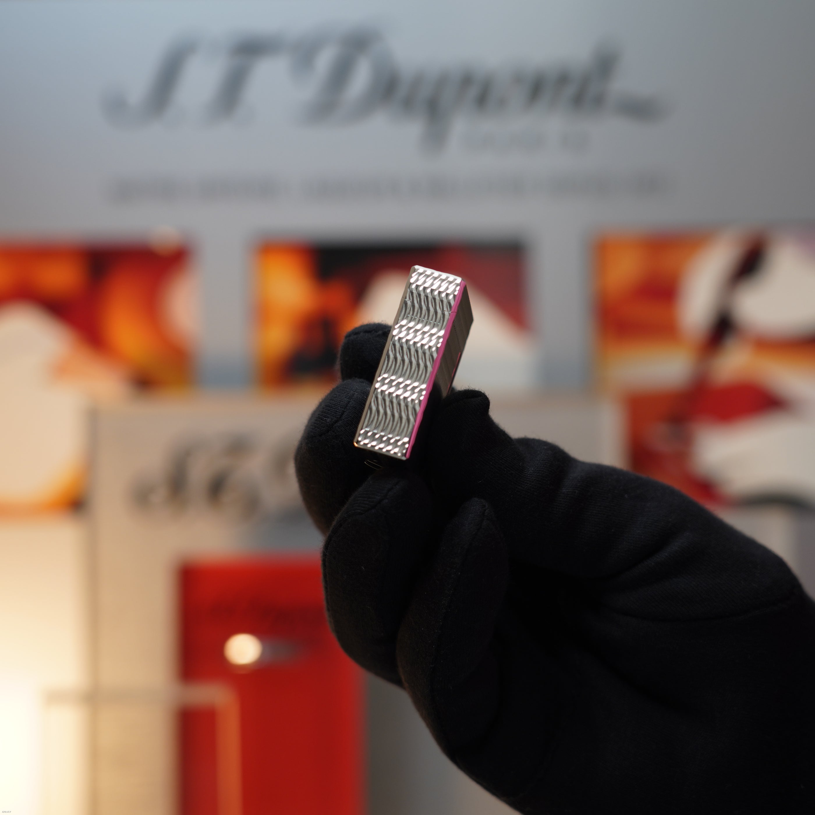 A gloved hand holds a Vintage 1990 Lucy Lighter S.T. Dupont Ligne 1 Small Size Rare 3D Wave Pattern Heavy Silver Finish lighter, with a blurred S.T. Dupont sign and colorful background visible behind it.