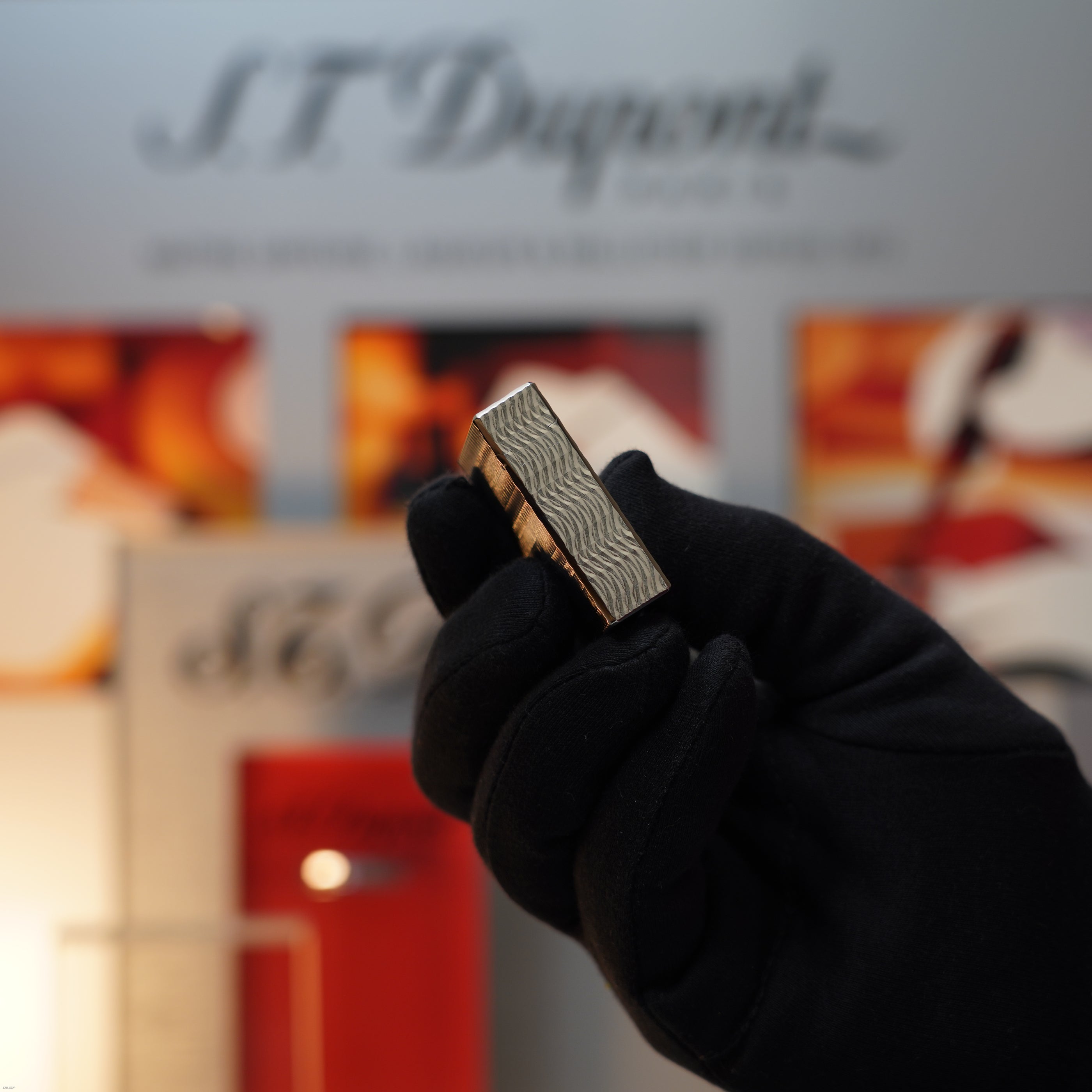 A gloved hand holds a Vintage 1990 Lucy Lighter S.T. Dupont Ligne 1 Small Size Rare 3D Wave Pattern Heavy Silver Finish lighter in front of a blurred S.T. Dupont display.