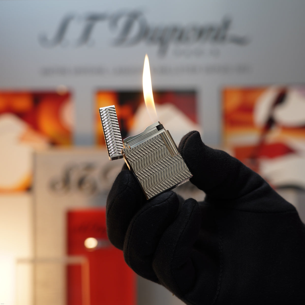 A gloved hand holds and lights a Vintage 1990 Lucy Lighter S.T. Dupont Ligne 1 Small Size 3D Wave Pattern Heavy Silver Finish Lighter, with blurred store displays in the background.