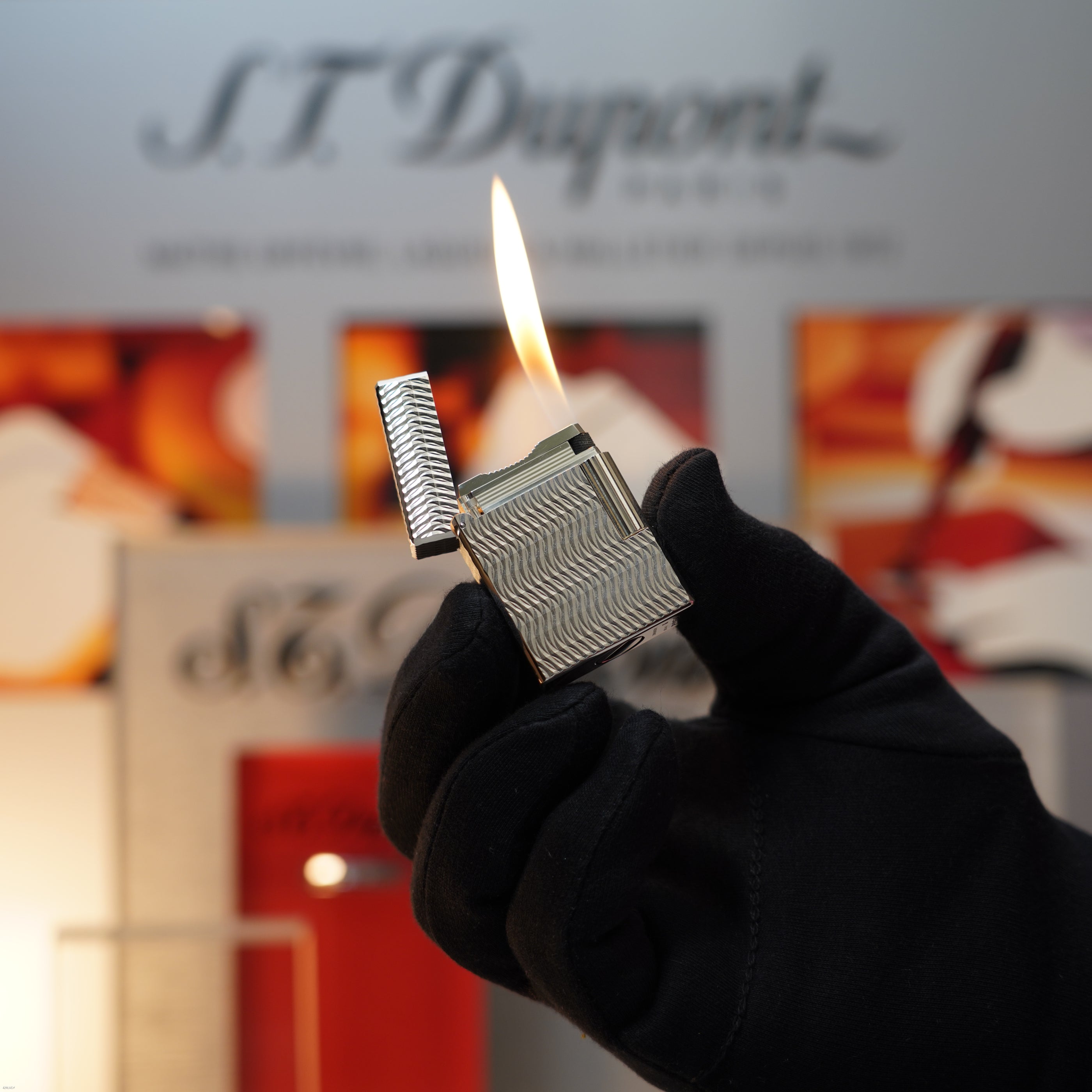 A gloved hand holds a Vintage 1990 Lucy Lighter S.T. Dupont Ligne 1 Small Size Rare 3D Wave Pattern Heavy Silver Finish Lighter, standing out against blurred branding and displays in the background.