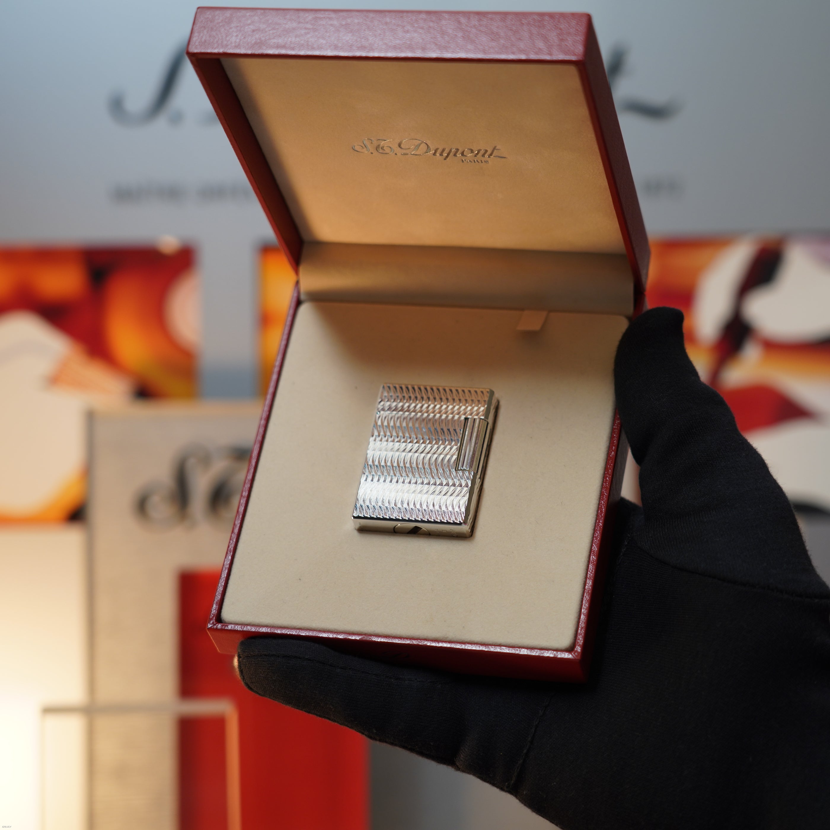 A gloved hand holds an open red box displaying a Vintage 1990 S.T. Dupont Ligne 1 Small Size Rare 3D Wave Pattern Heavy Silver Finish Lighter by Lucy Lighter, set against a blurred background.