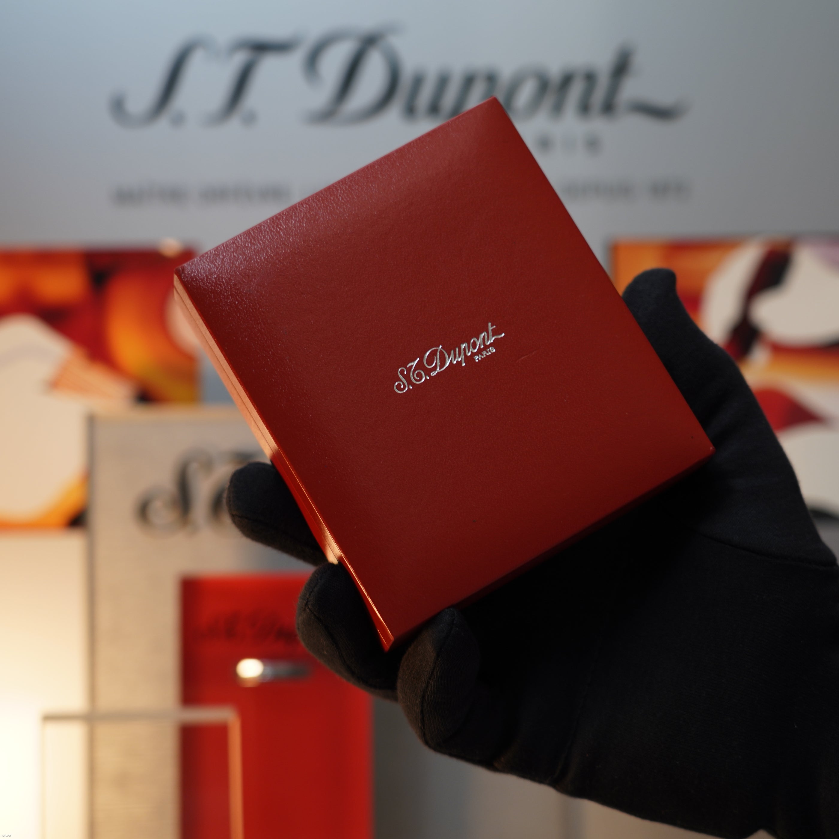 A gloved hand holds a red S.T. Dupont Paris box with silver lettering, likely containing a Lucy Lighter Vintage 1990 S.T. Dupont Ligne 1 Small Size Rare 3D Wave Pattern Heavy Silver Finish Lighter, against a blurred display backdrop.