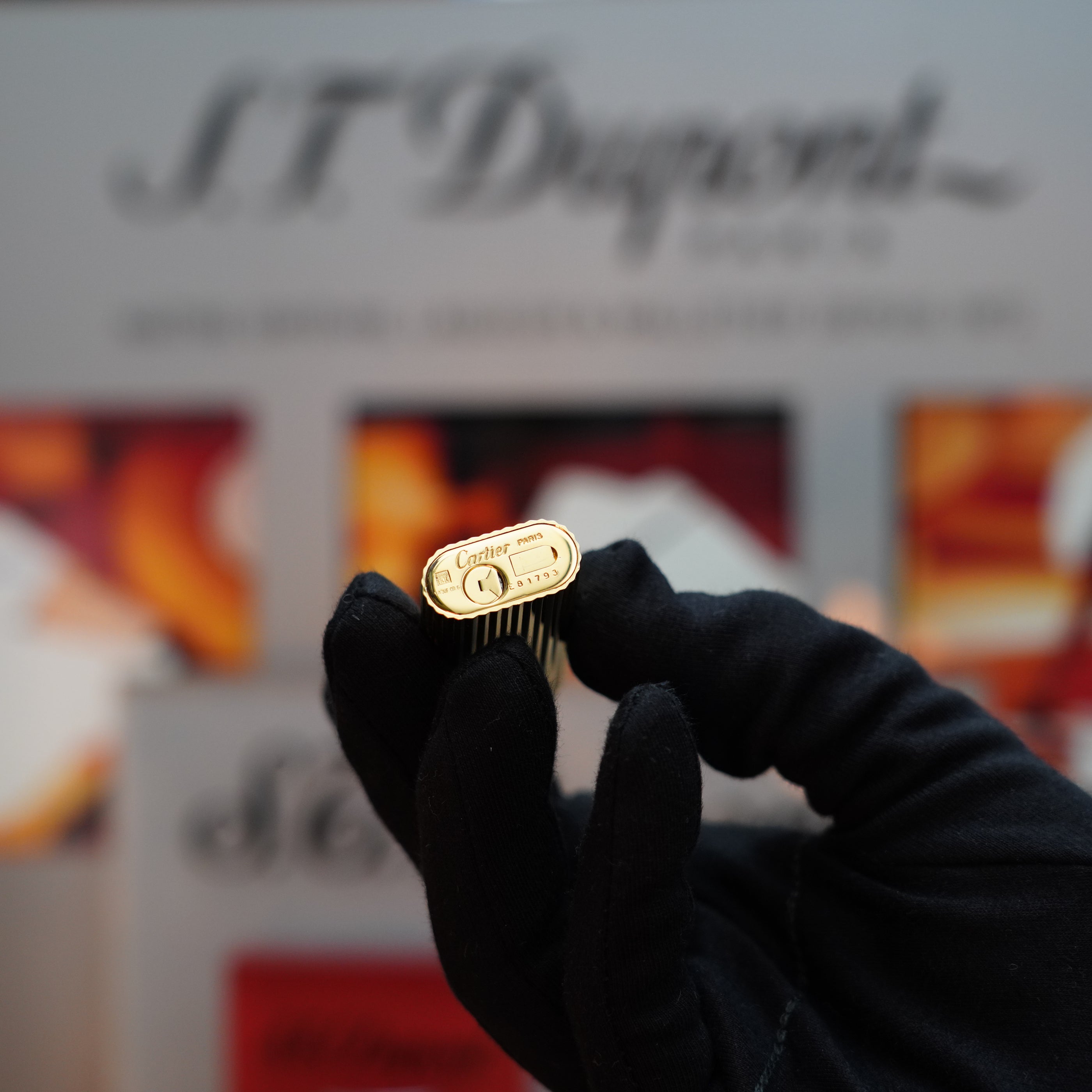 In the background, a gloved hand presents a gold Cartier pen cap beside an S.T. Dupont display, evoking the refined elegance of Cartier's Vintage 1980 Vertical Line Le Must Lighter with its 24k Gold Finish.