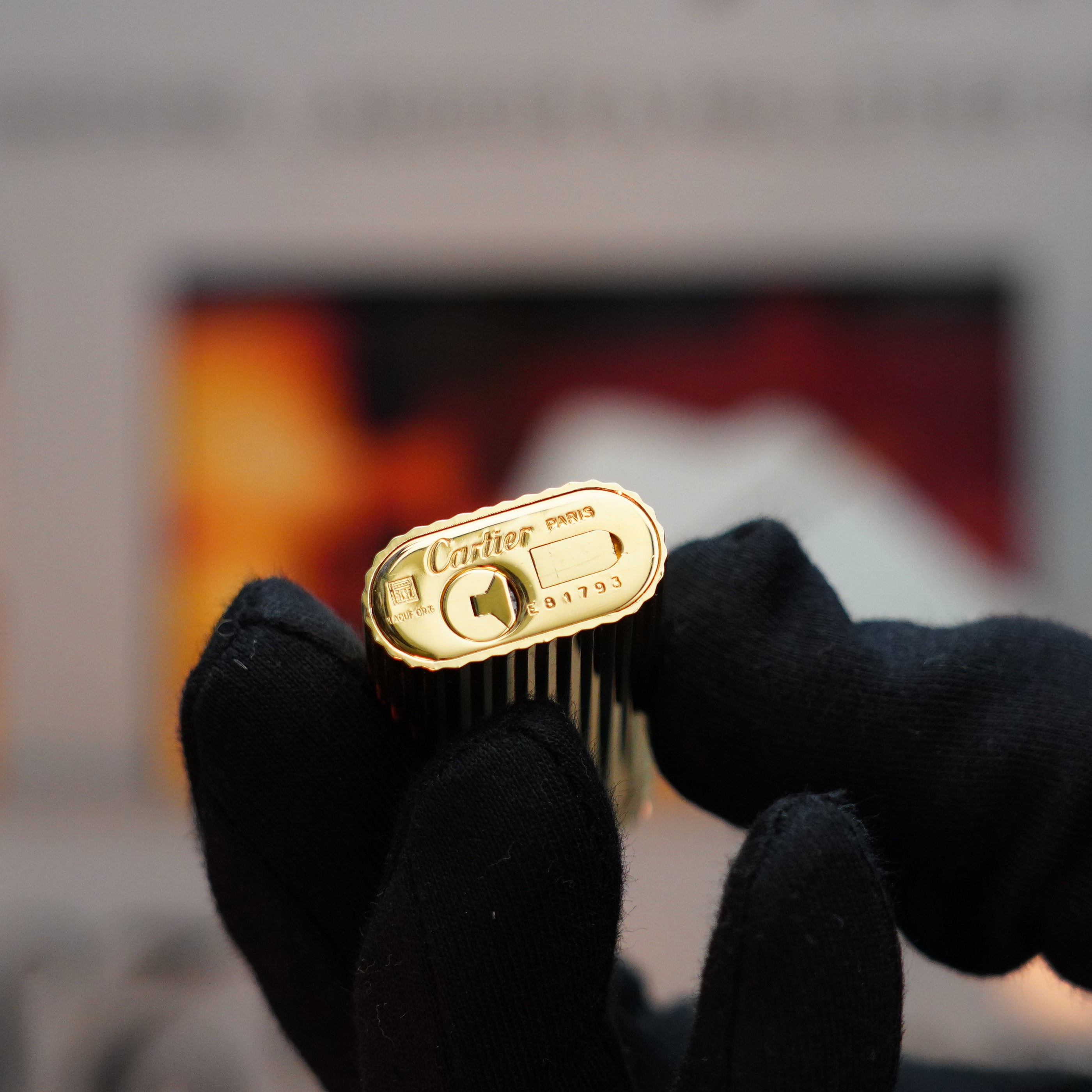 A gloved hand holds a small Cartier lighter from the Vintage 1980 Le Must series, viewed from the bottom, its 24k gold finish gleaming softly.