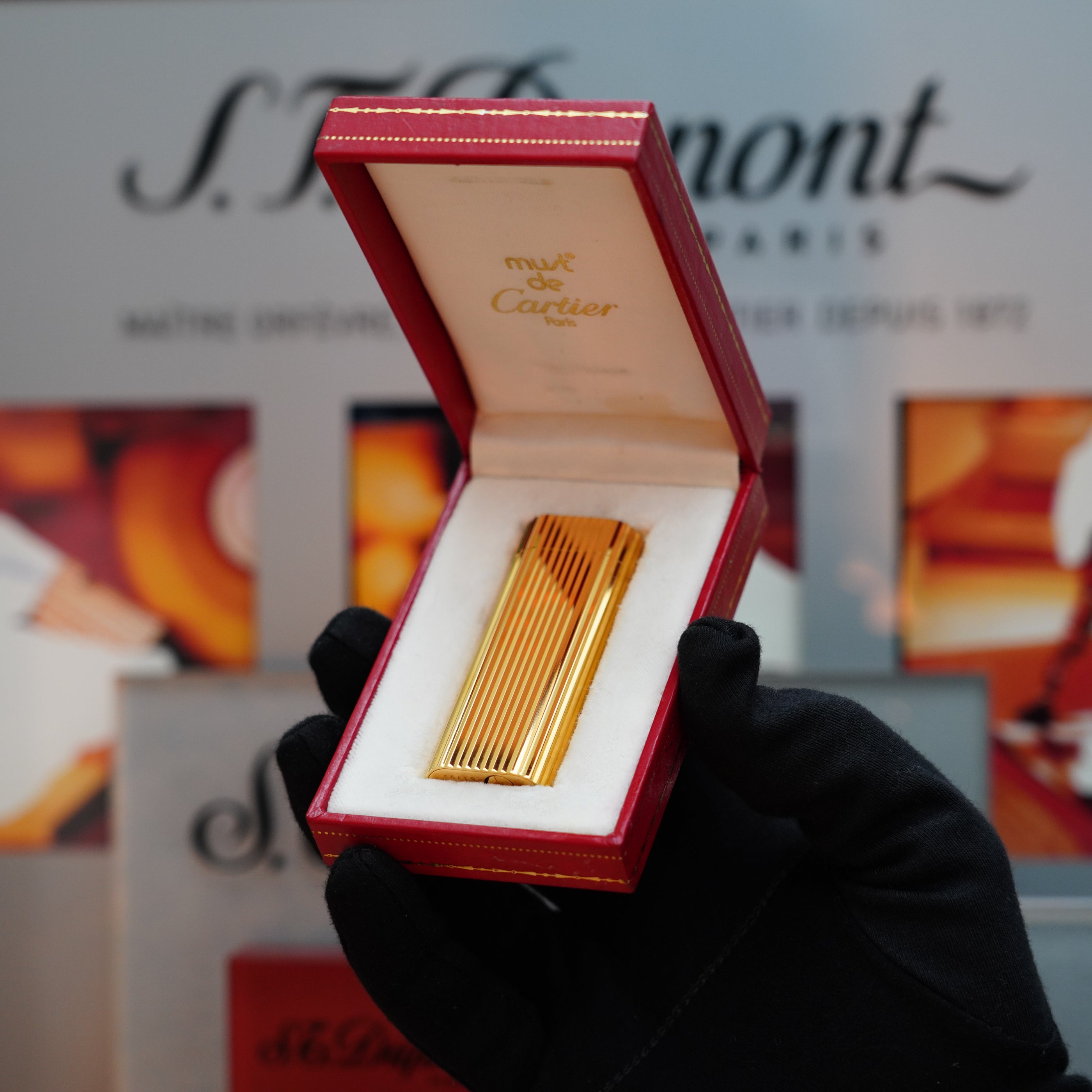 A gloved hand presents an open red box, showcasing the gold rectangular Vintage 1980 Cartier Vertical line 24k Gold Finish Le Must Lighter. The lid displays "must de Cartier," suggesting the sophistication of the Le Must Series. In the background, blurred text and designs emphasize this vintage Cartie lighter's timeless allure.