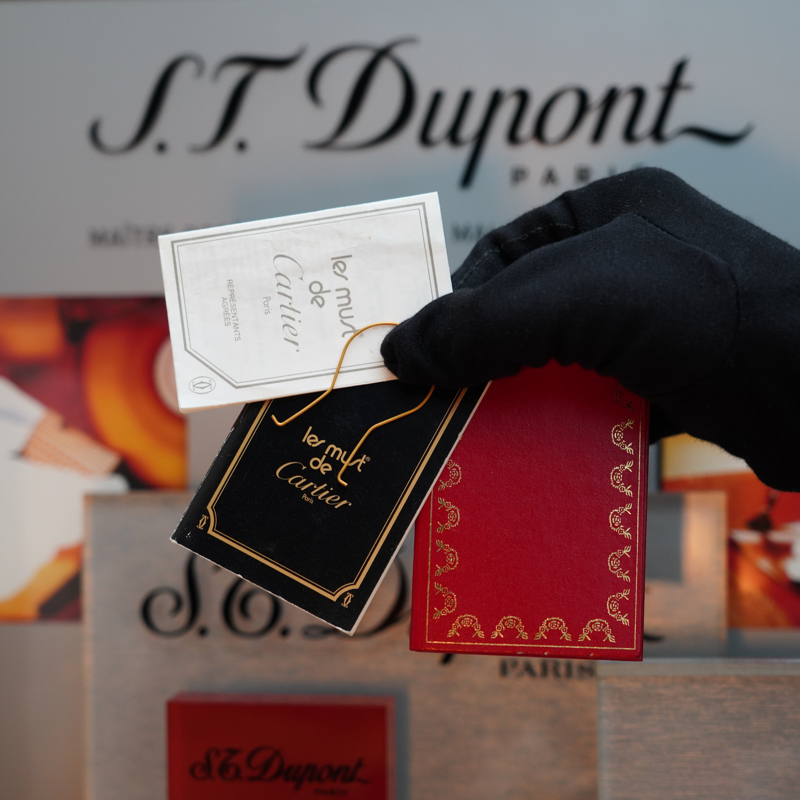 A gloved hand holds two Cartier perfume cards from the Le Must Series in front of a display featuring S.T. Dupont branding, echoing the sophistication of a Vintage 1980 Cartier Vertical Line 24k Gold Finish Le Must Lighter.