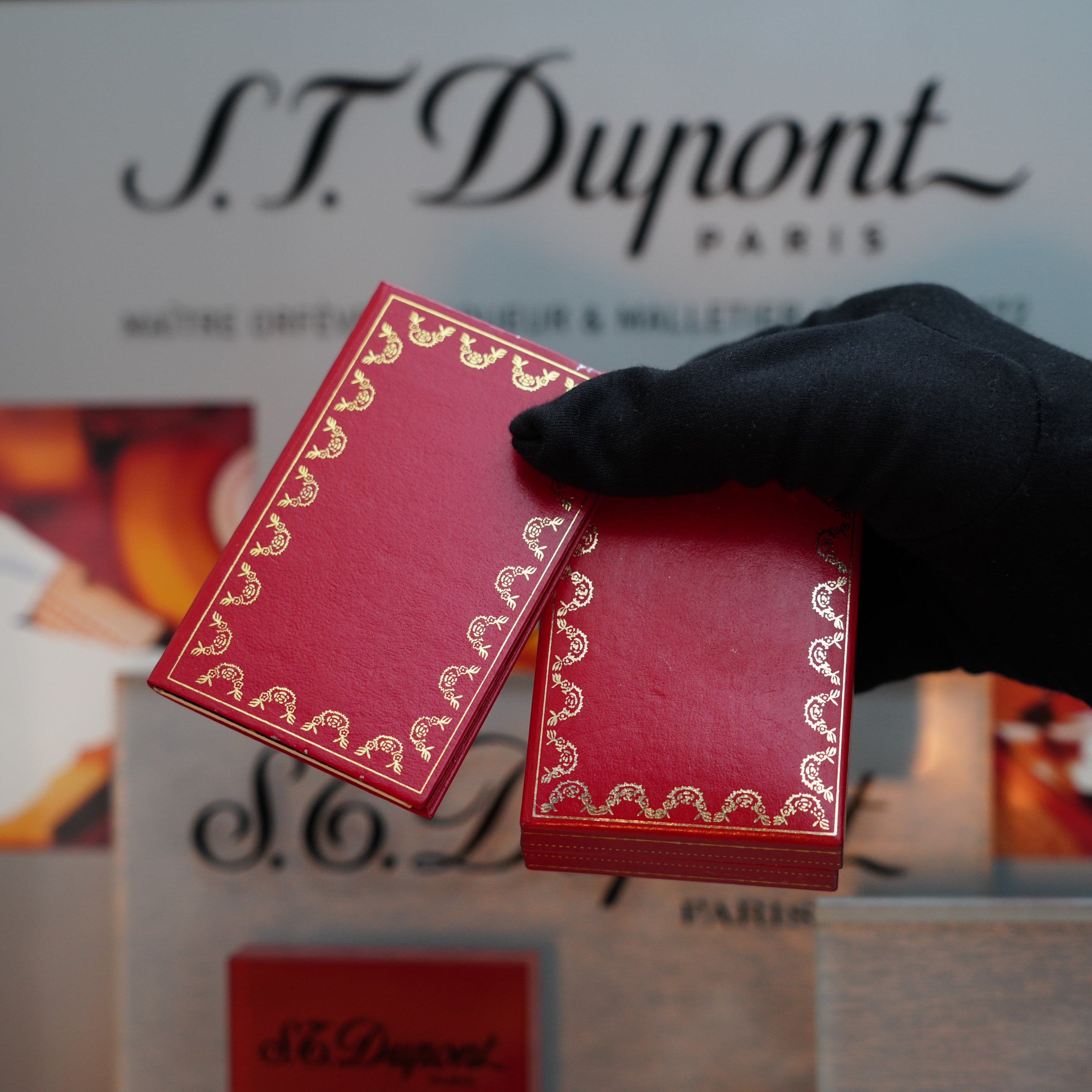 A gloved hand presents two red items with gold detailing from the S.T. Dupont Le Must series against a branded backdrop, capturing the elegance of a Vintage 1980 Cartier Vertical Line 24k Gold Finish Le Must Lighter.