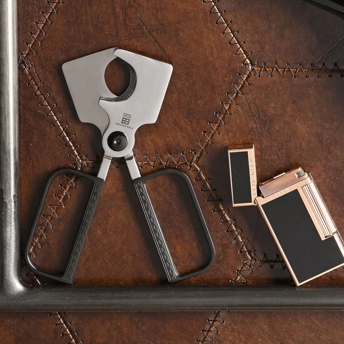 A silver and black cigar cutter tool is shown on a brown stitched leather surface next to two slim rectangular lighters with black and rose-gold finishes.