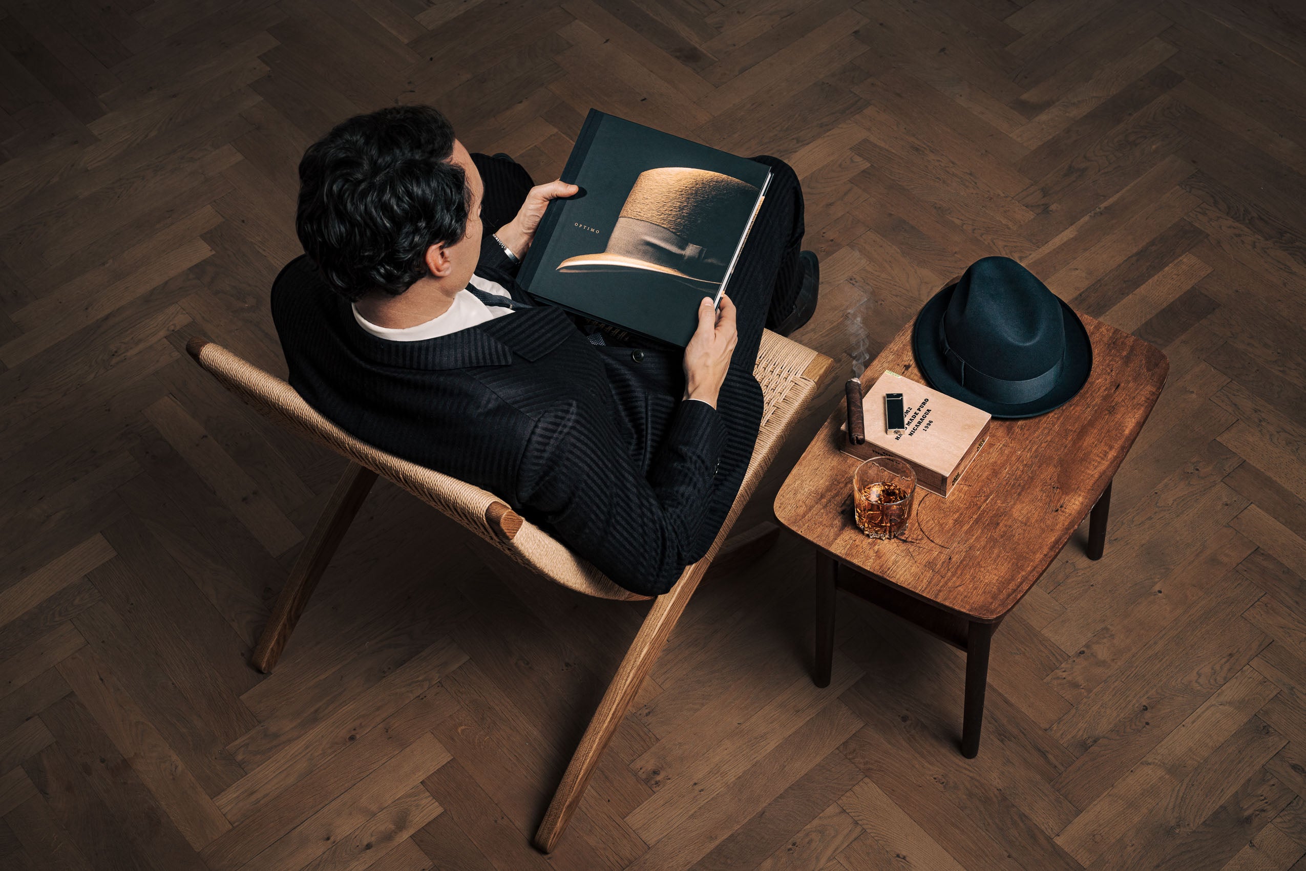 A man sitting in a chair reading the Optimo Hats: The Art of the Hatmaker book from KirbyAllison.com.