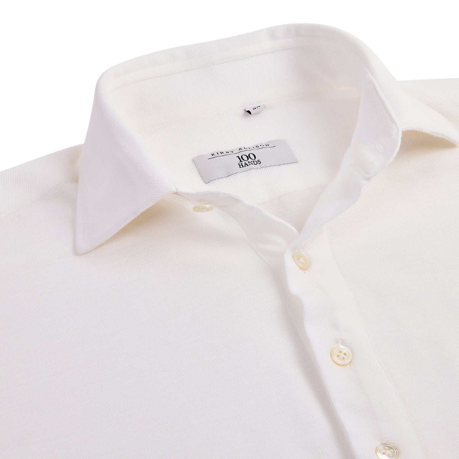 Close-up of a neatly folded white long sleeve knit polo with a crisp collar and buttons visible. The label inside reads "100 Hands," part of the exclusive 100 Hands x Kirby Allison collection available on KirbyAllison.com.