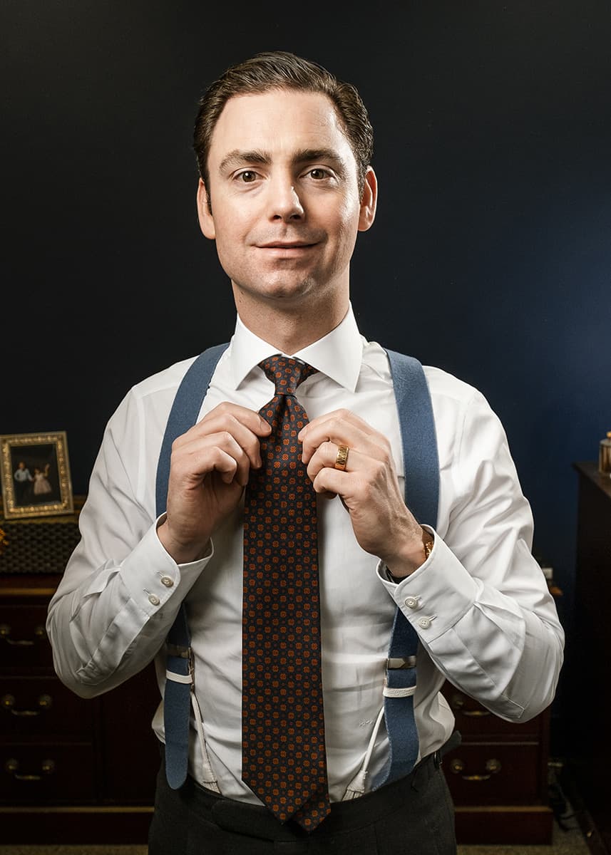 An image of Kirby Allison standing up, in white dress shirt and blue suspenders, and adjusting his patterned tie.