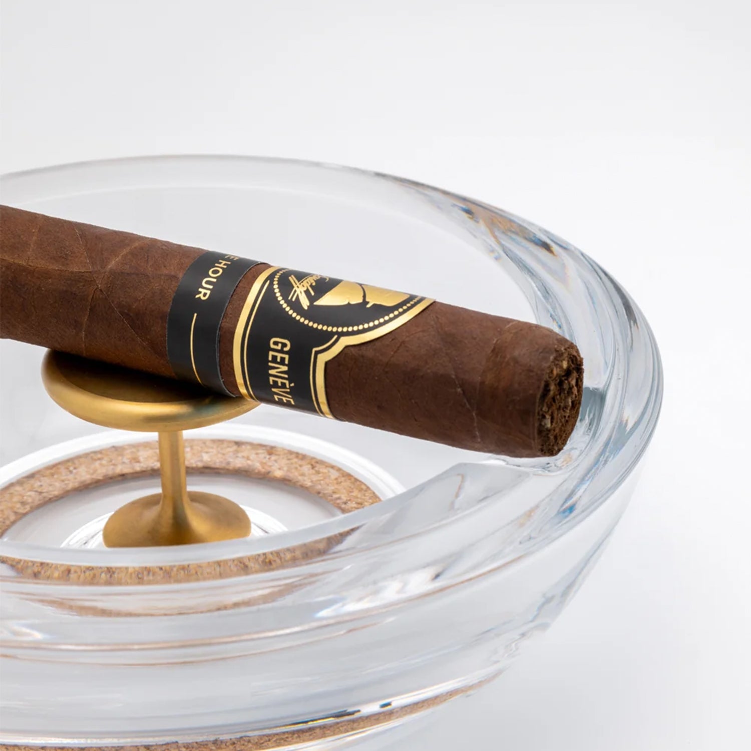 A single dark brown cigar resting across a clear glass ashtray with a gold metal stand on a clean white background.