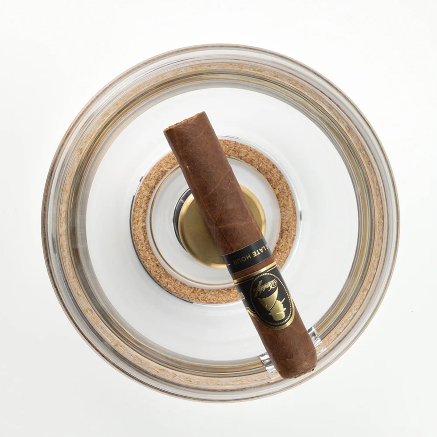 A single brown cigar with a black and gold wrapper band, resting diagonally inside a clear glass ashtray with a cork insert on a white background.