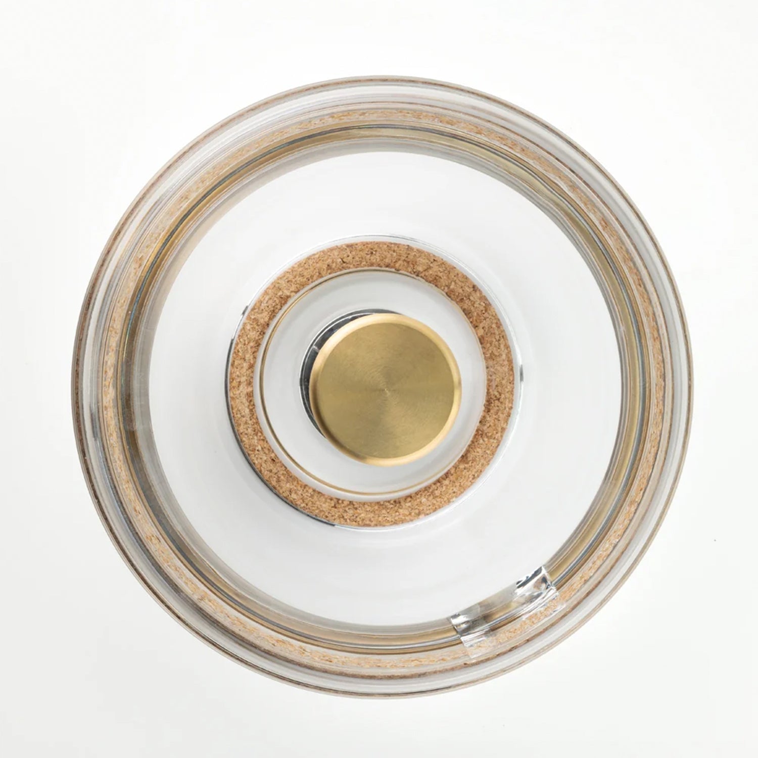Top-down view of a circular clear glass container or jar lid with a natural cork ring and a central brushed gold metal button on a white background.