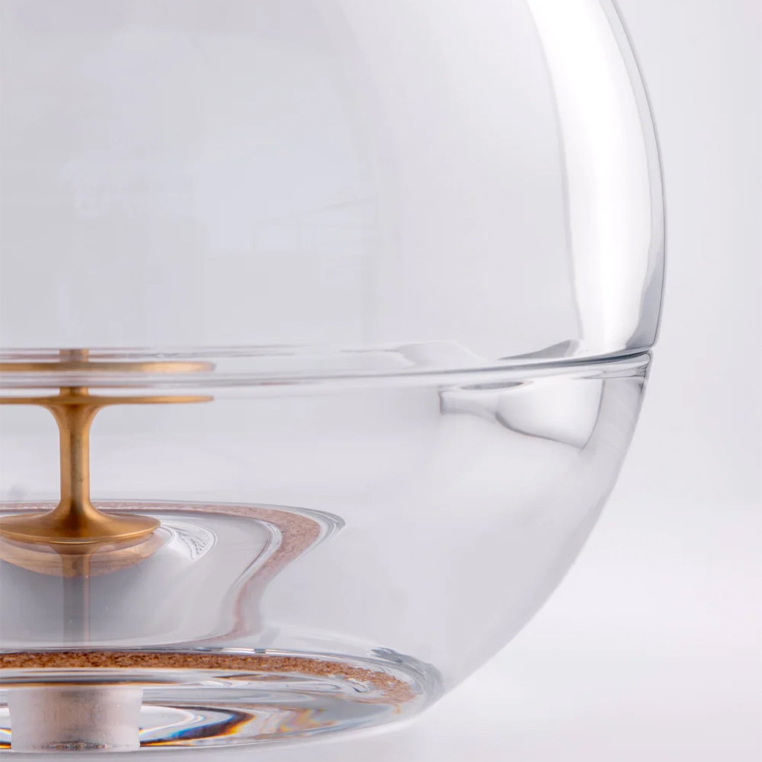 Close-up of a transparent glass spherical ashtray with a metallic gold central stem and a thin copper-toned ring near the bottom, photographed on a clean white background.